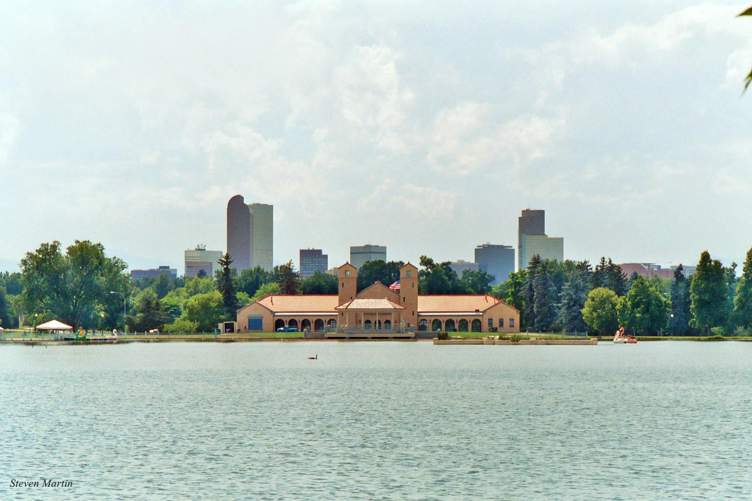 A large building across a lake with a distant city skyline under a cloudy sky.