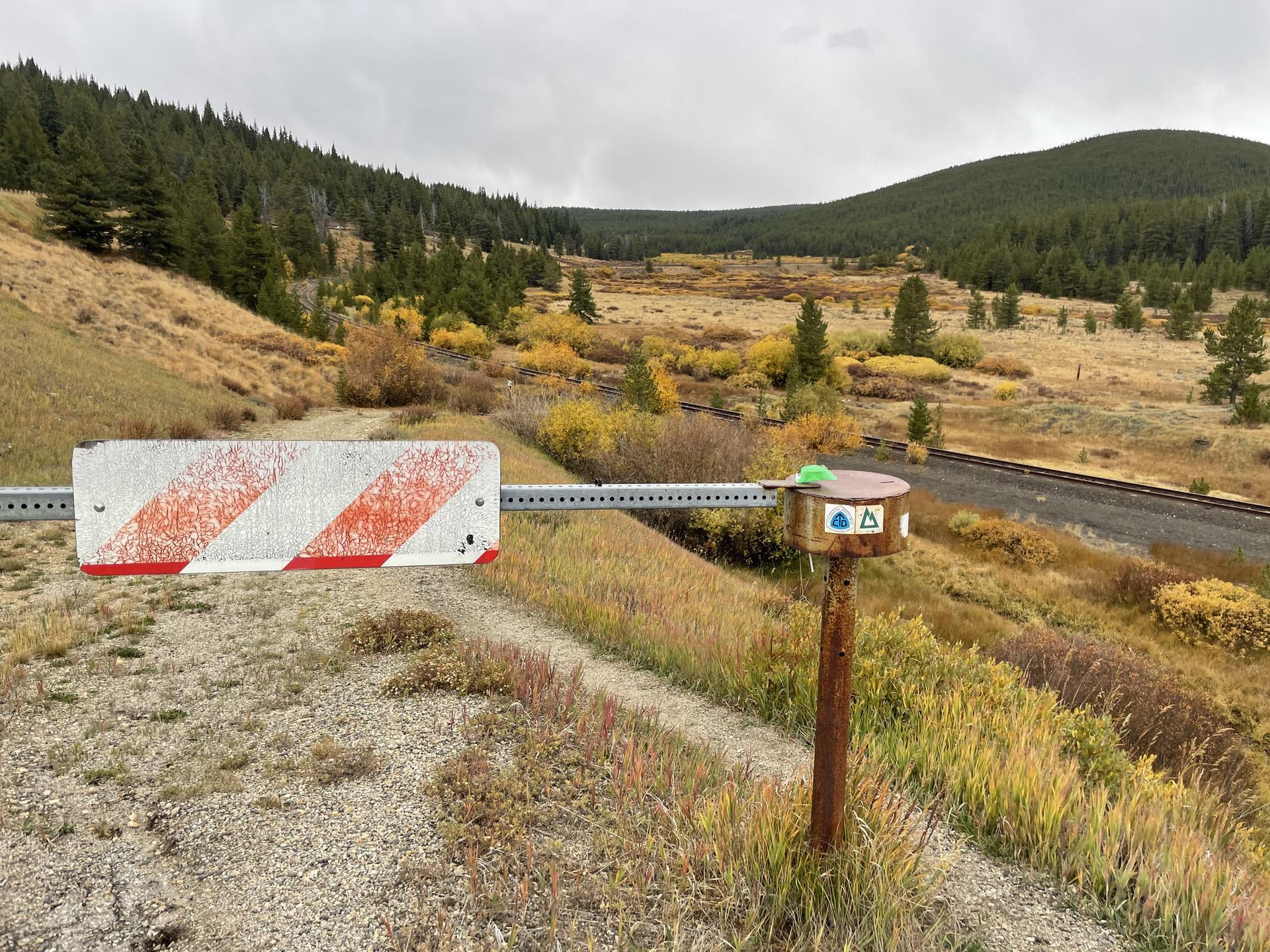 A red and white barrier blocks a dirt path next to a signpost.