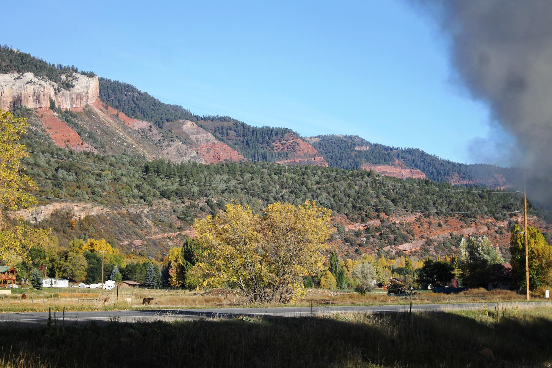 Bright mountain scene with autumn foliage, a road, and a large smoke plume.