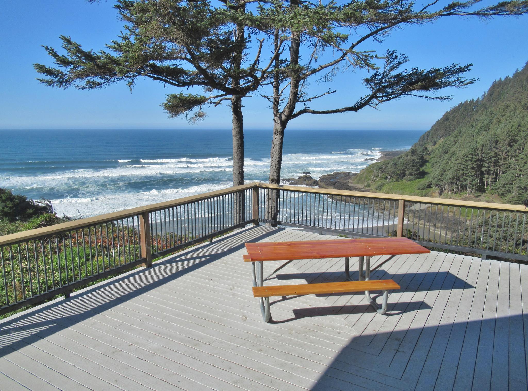 Wooden deck with picnic table overlooking a sunny ocean and forested coast.