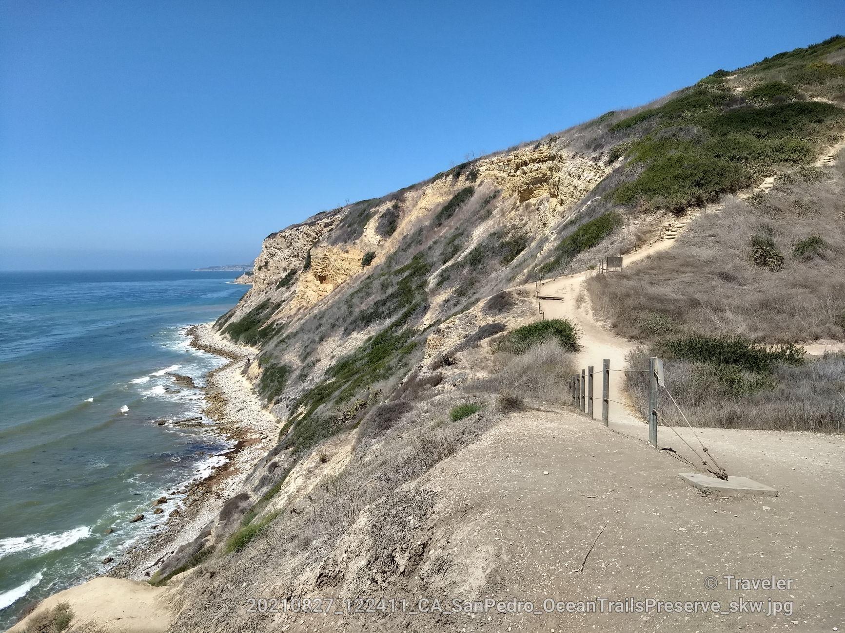 A winding dirt trail along steep, rocky coastal cliffs under a clear sky.