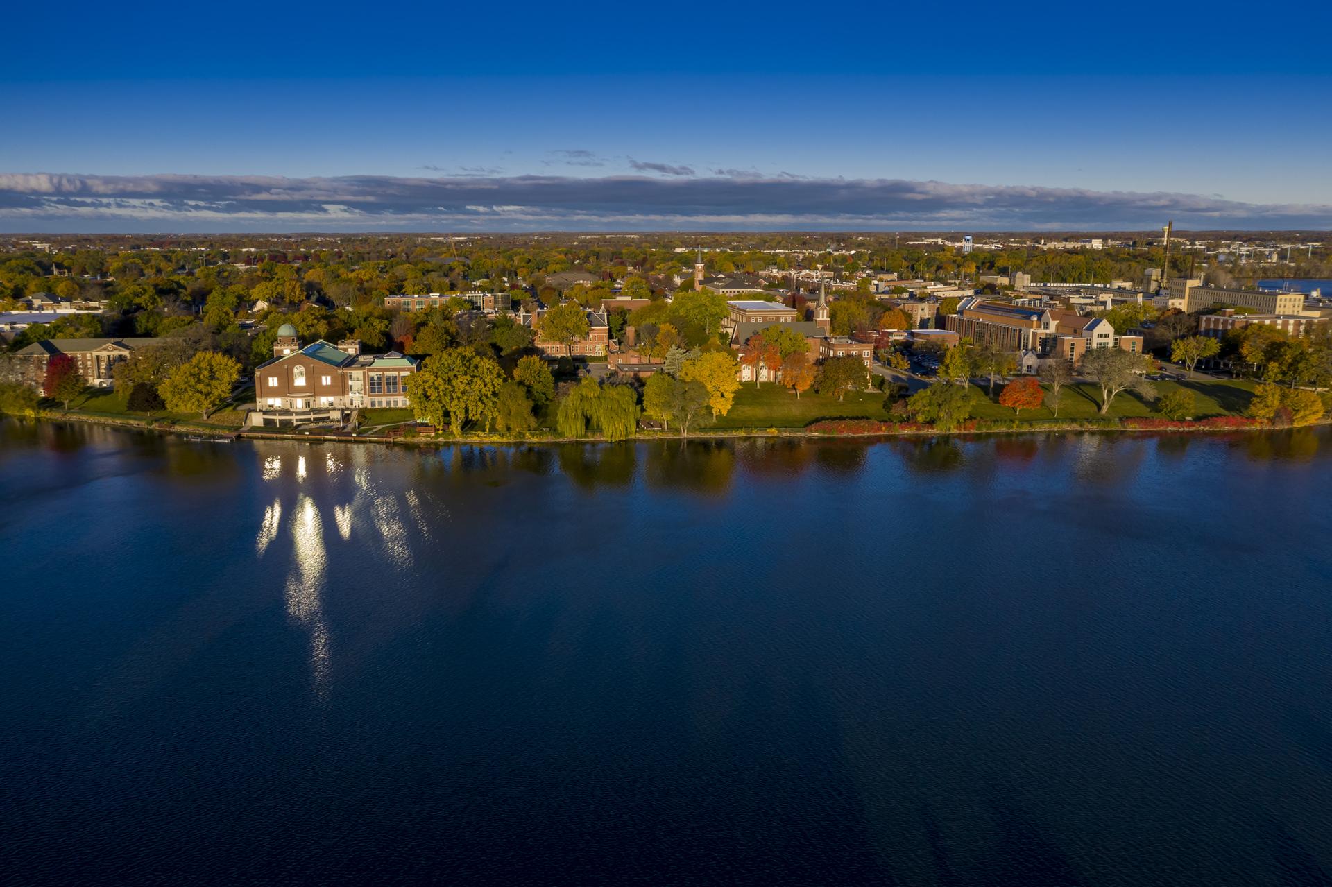 Aerial view of buildings and trees lining a dark blue lake under a clear sky.