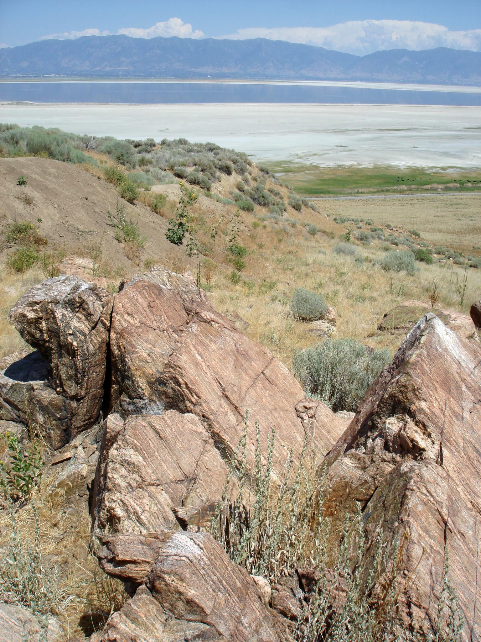 Large textured rocks in foreground, dry hillside, and a vast lake under sunny skies.