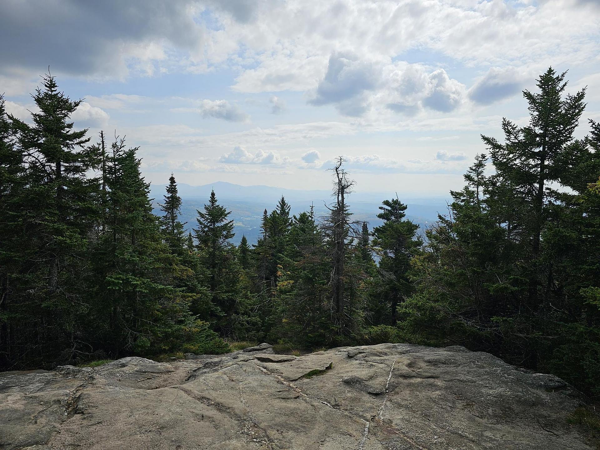Rocky overlook with evergreen trees and distant forested mountains under a cloudy sky.