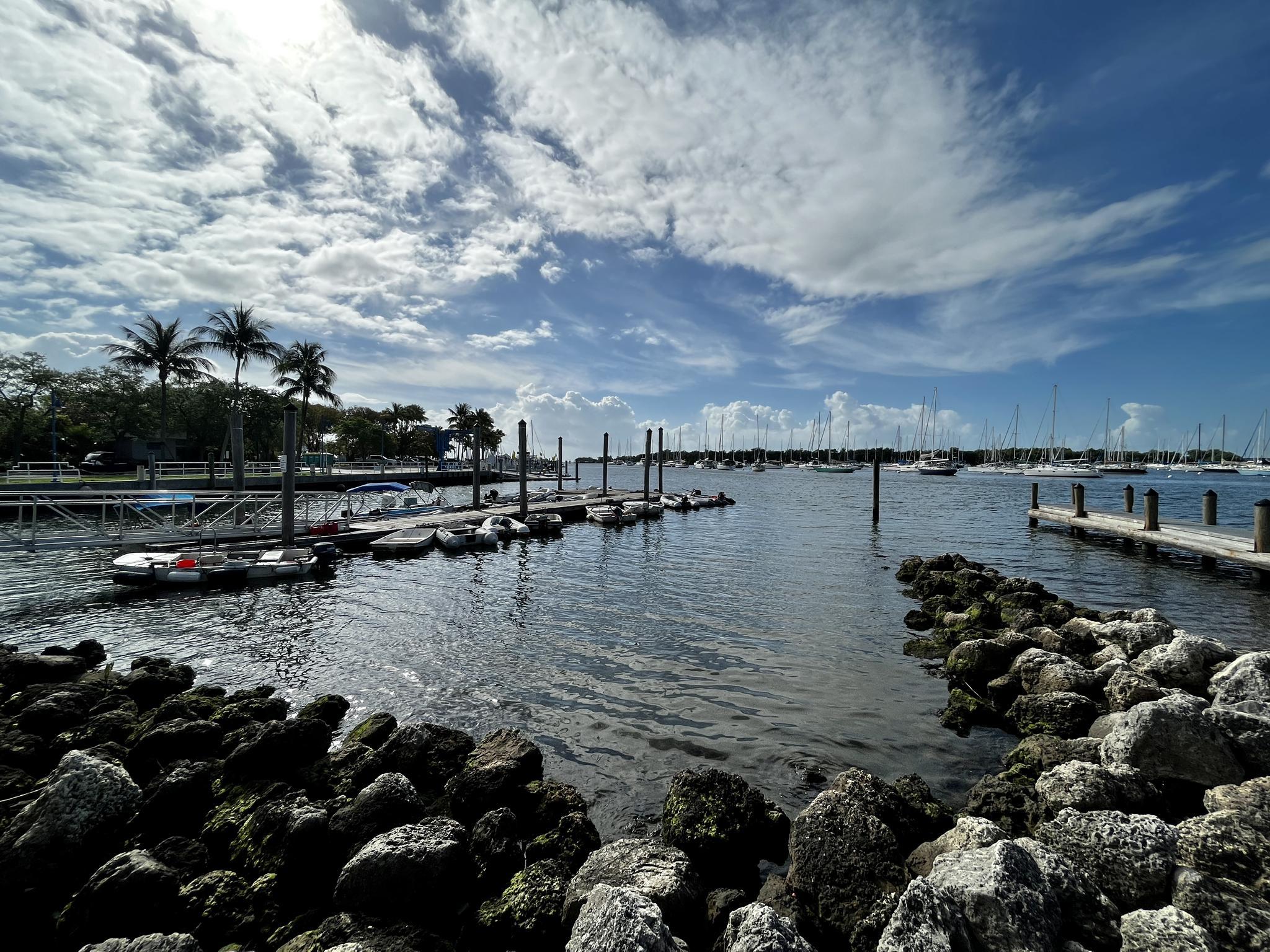 Sunny marina with boats docked, rocky shore in foreground, under a cloudy sky.