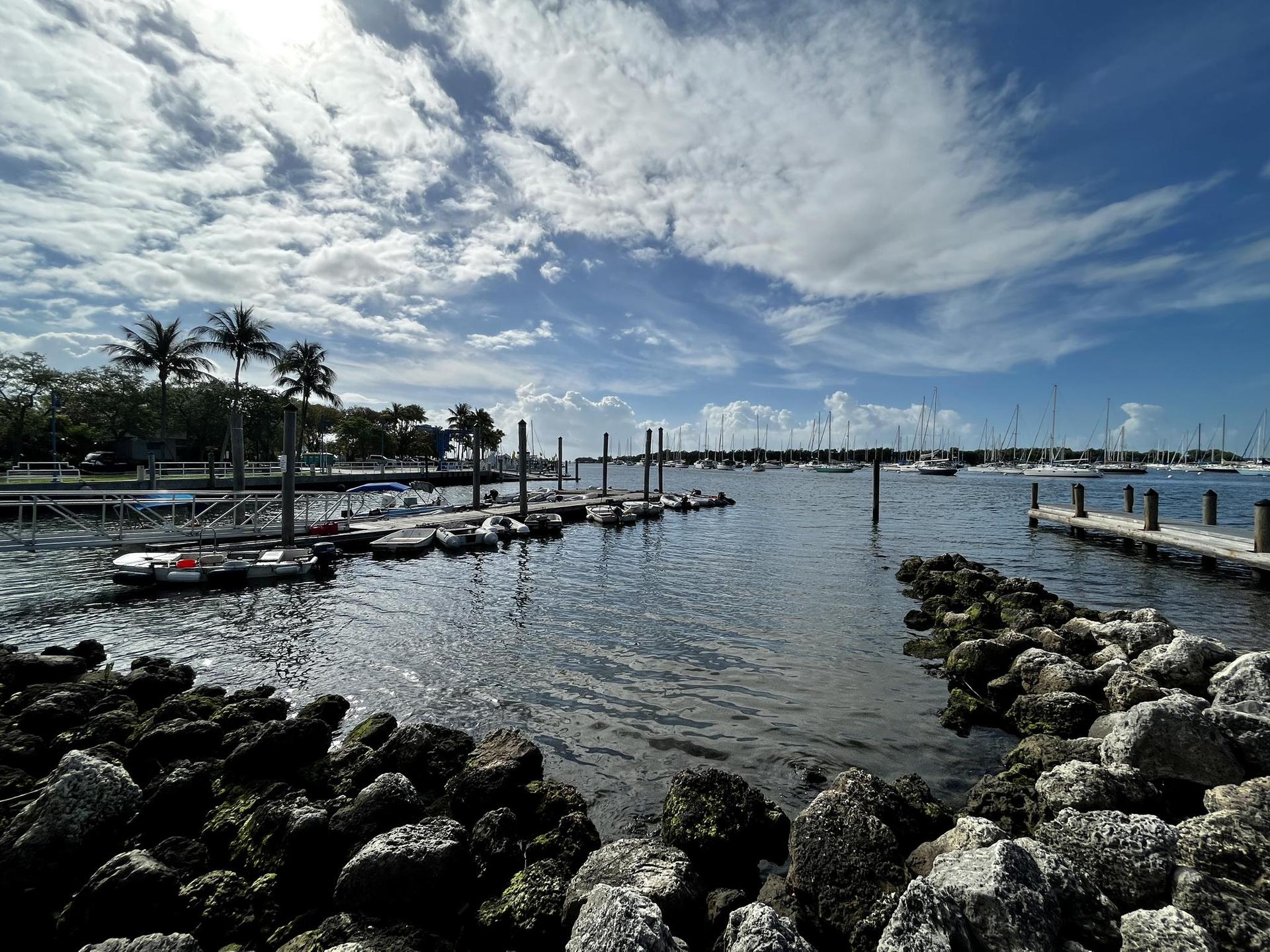 Sunny marina with boats docked, rocky shore in foreground, under a cloudy sky.