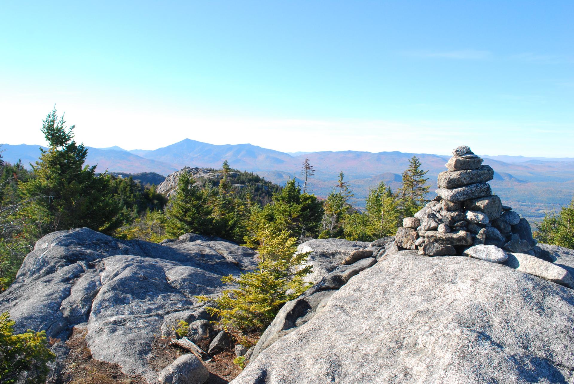 A rock cairn on a sunny mountain summit overlooking distant peaks.