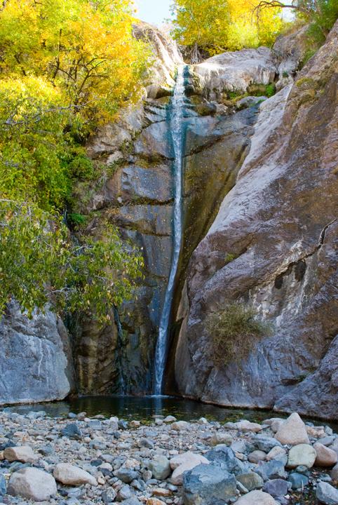 A tall, narrow waterfall cascades into a rocky pool, surrounded by autumn trees.