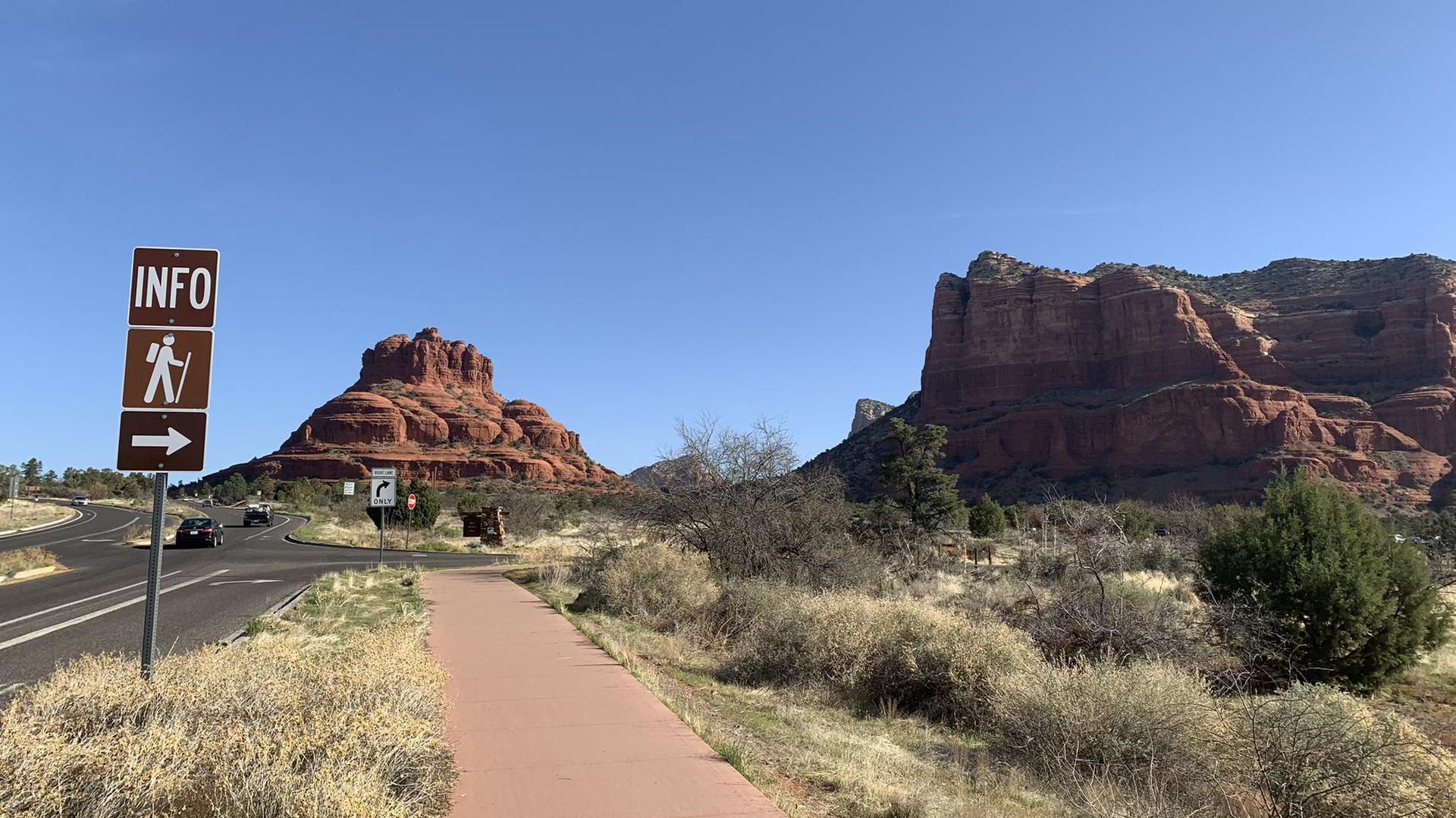 Road and sidewalk leading towards red rock formations under a clear blue sky.