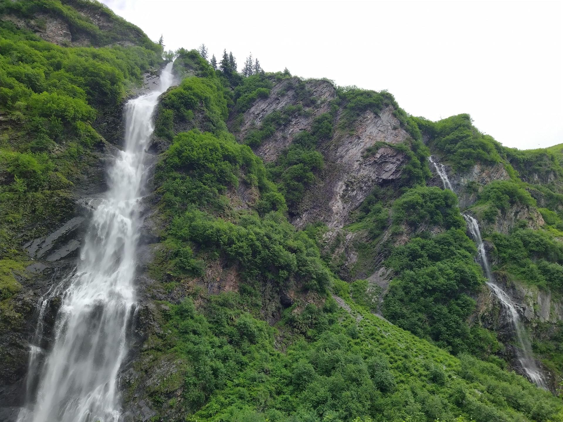 Multiple waterfalls flow down a steep, green, rocky mountainside on an overcast day.