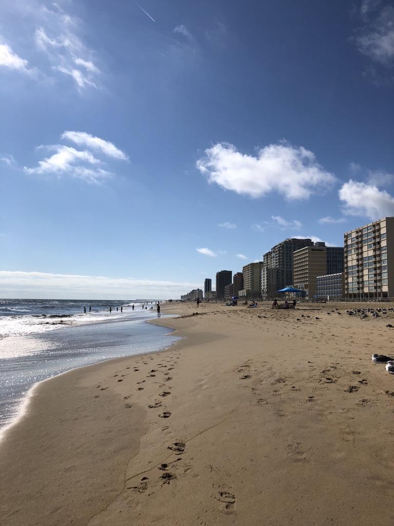 A sunny beach with footprints, ocean waves, and tall buildings under a blue sky.