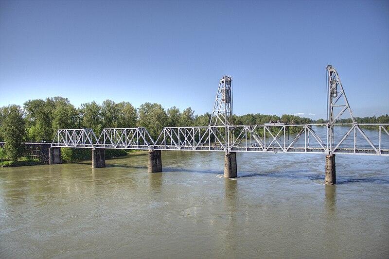 A long metal truss bridge spans a wide river under a clear blue sky.