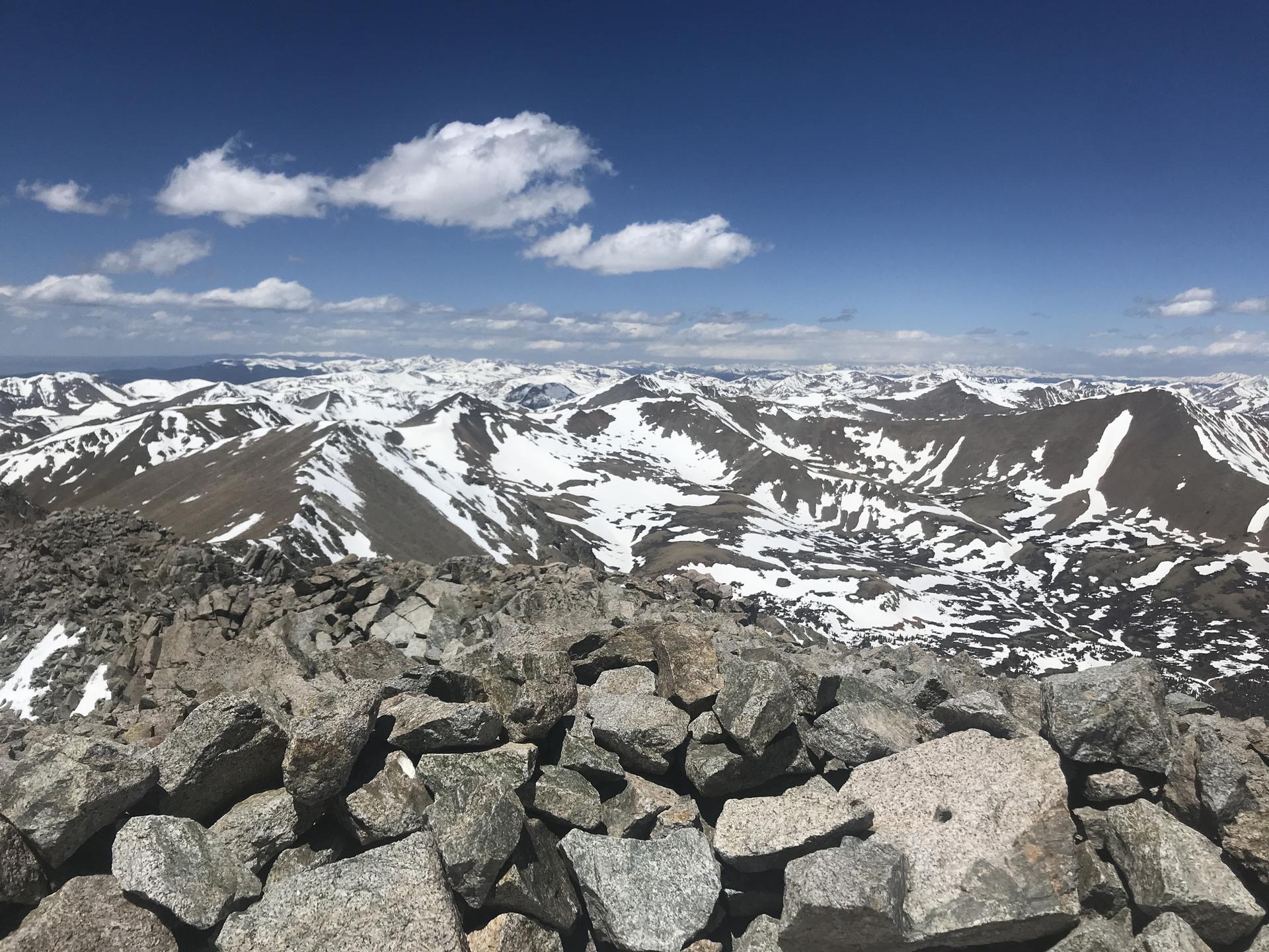 Vast snowy mountain peaks under a bright blue sky with a rocky foreground.