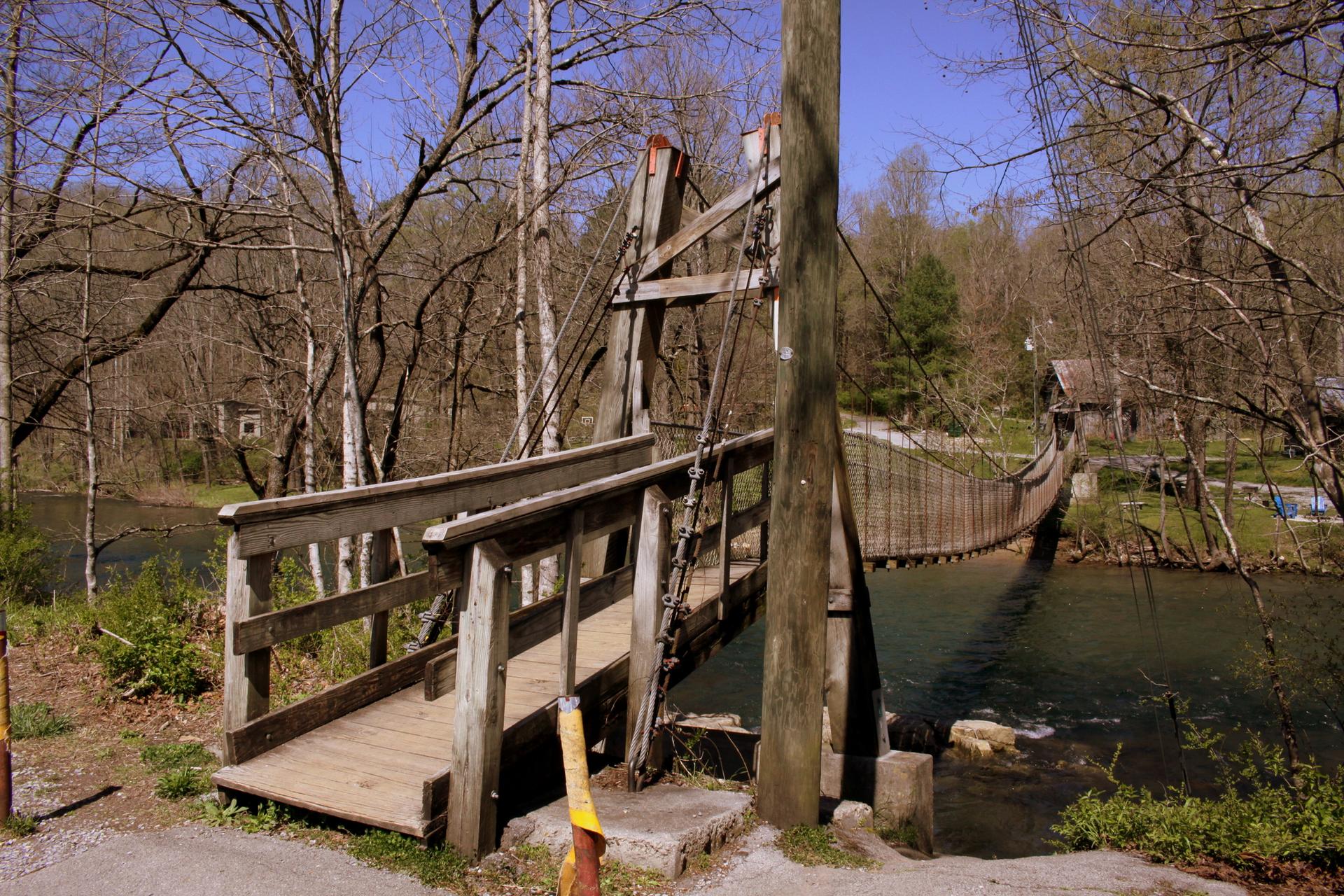 Wooden suspension bridge over a river with bare trees under bright daylight.
