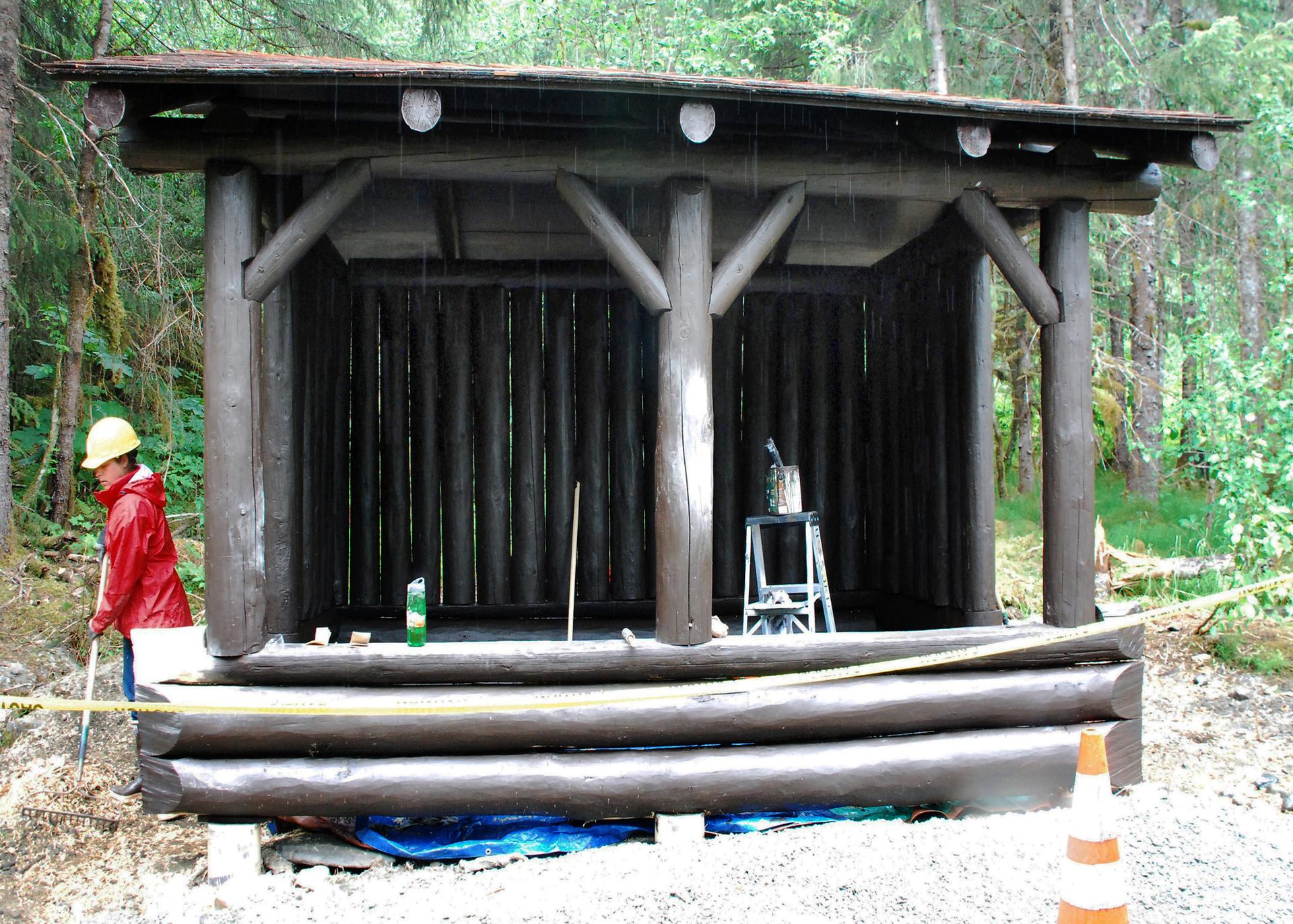 Person in red jacket and hard hat near a rustic wooden shelter in a forest.