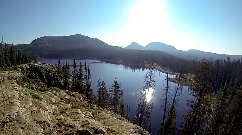 Sunny lake surrounded by mountains and pine trees, with bright sun glare.
