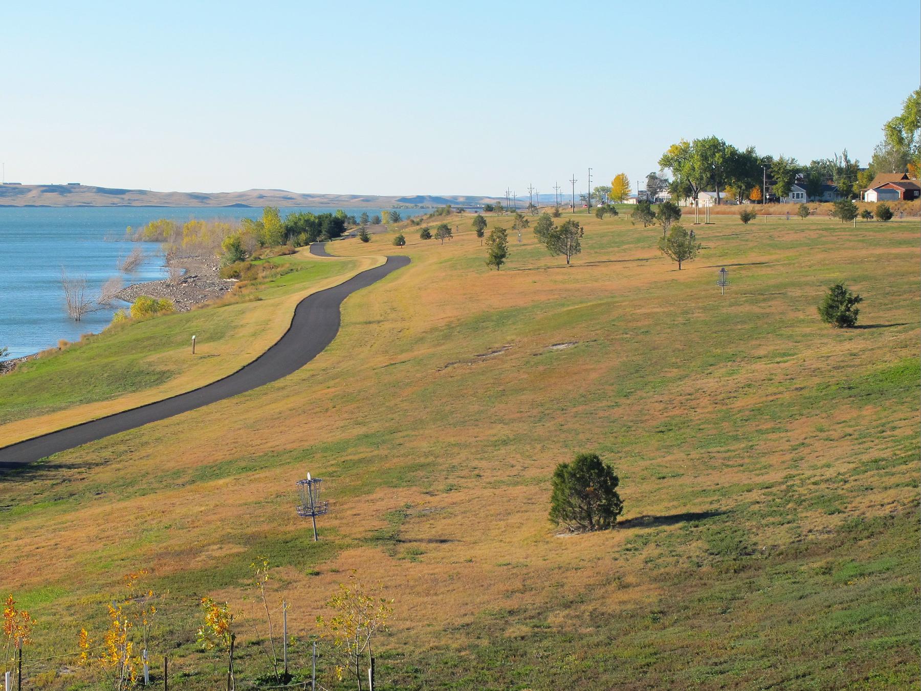A winding paved path along a sunny, grassy hillside next to blue water.