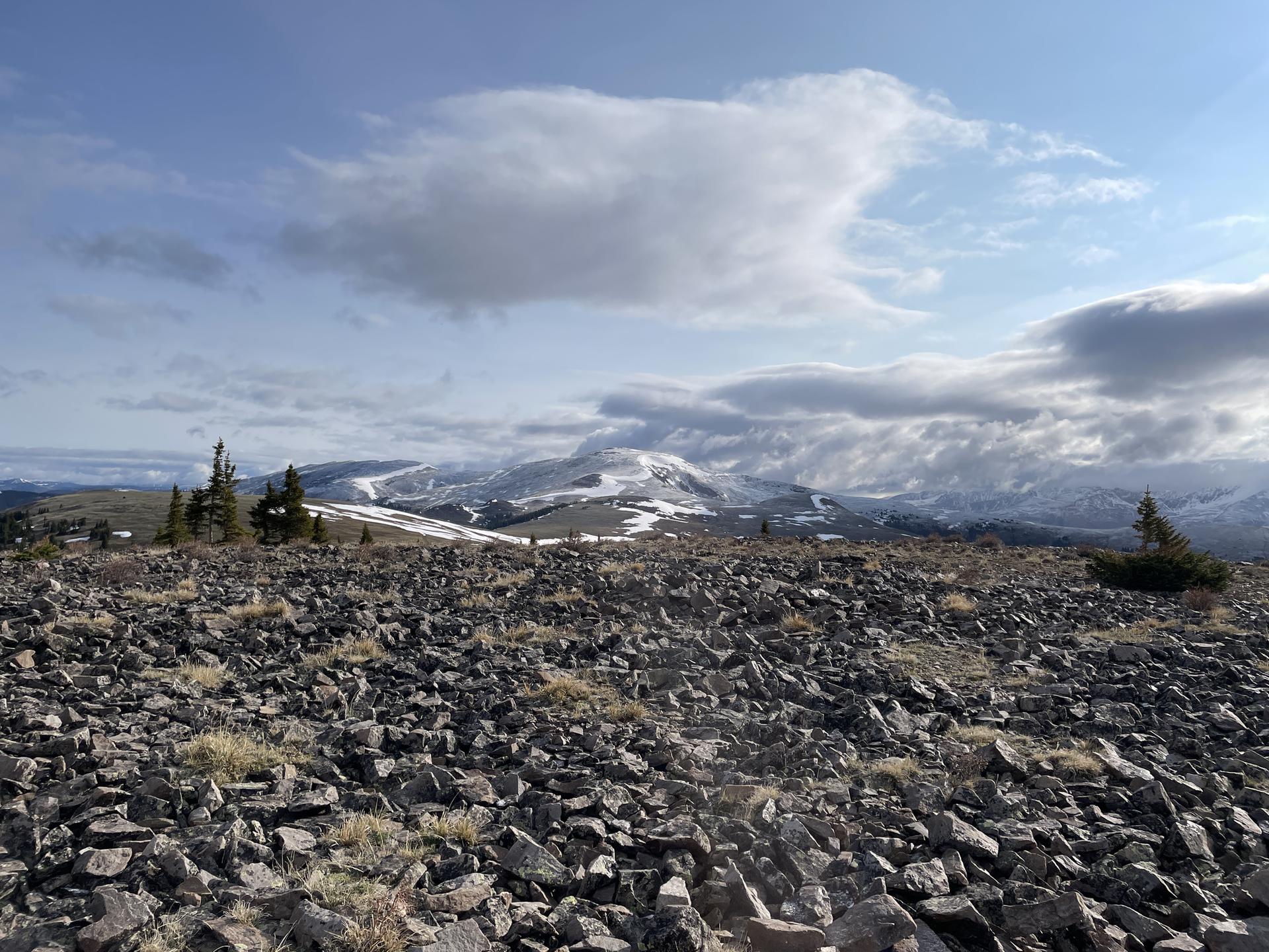 A vast rocky foreground overlooks distant snow-capped mountains under a cloudy sky.