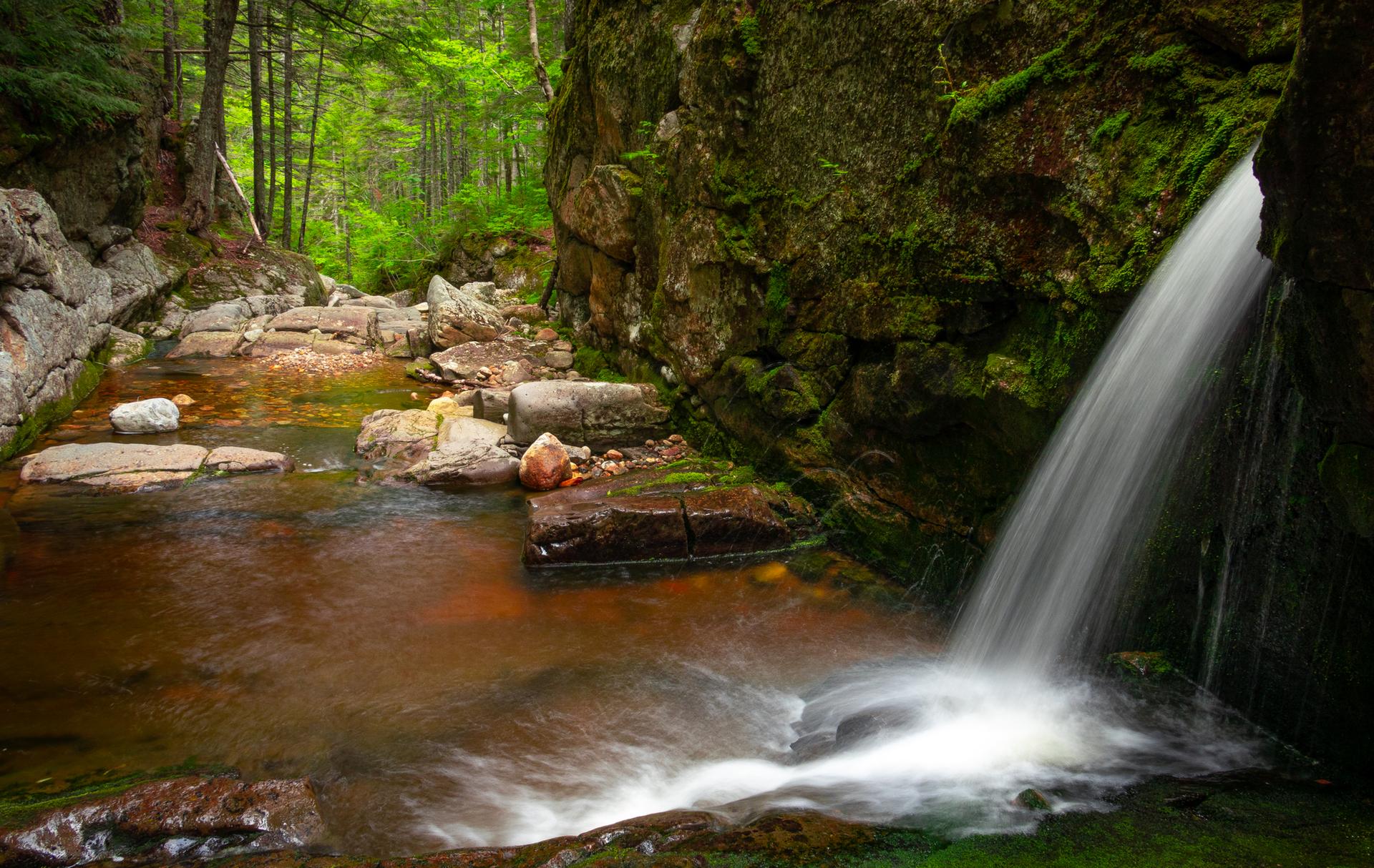 Waterfall plunging into a rocky, reddish stream in a shaded, mossy forest.