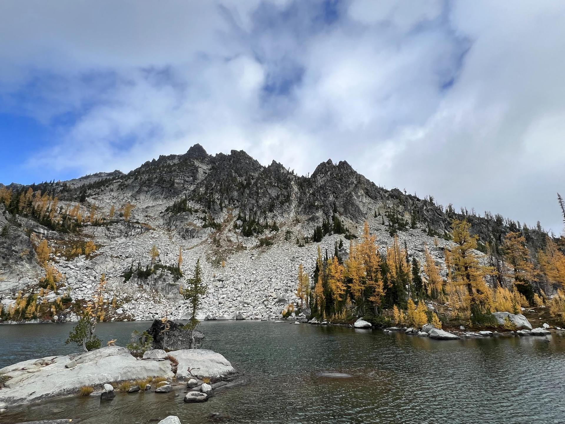 A lake with rocky shores, golden trees, and a rugged mountain under clouds.