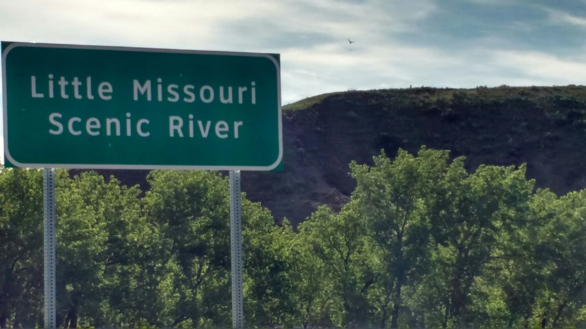 A green rectangular sign above green trees and a dark hill under a cloudy sky. - United States Bicycle Route 30 (North Dakota)