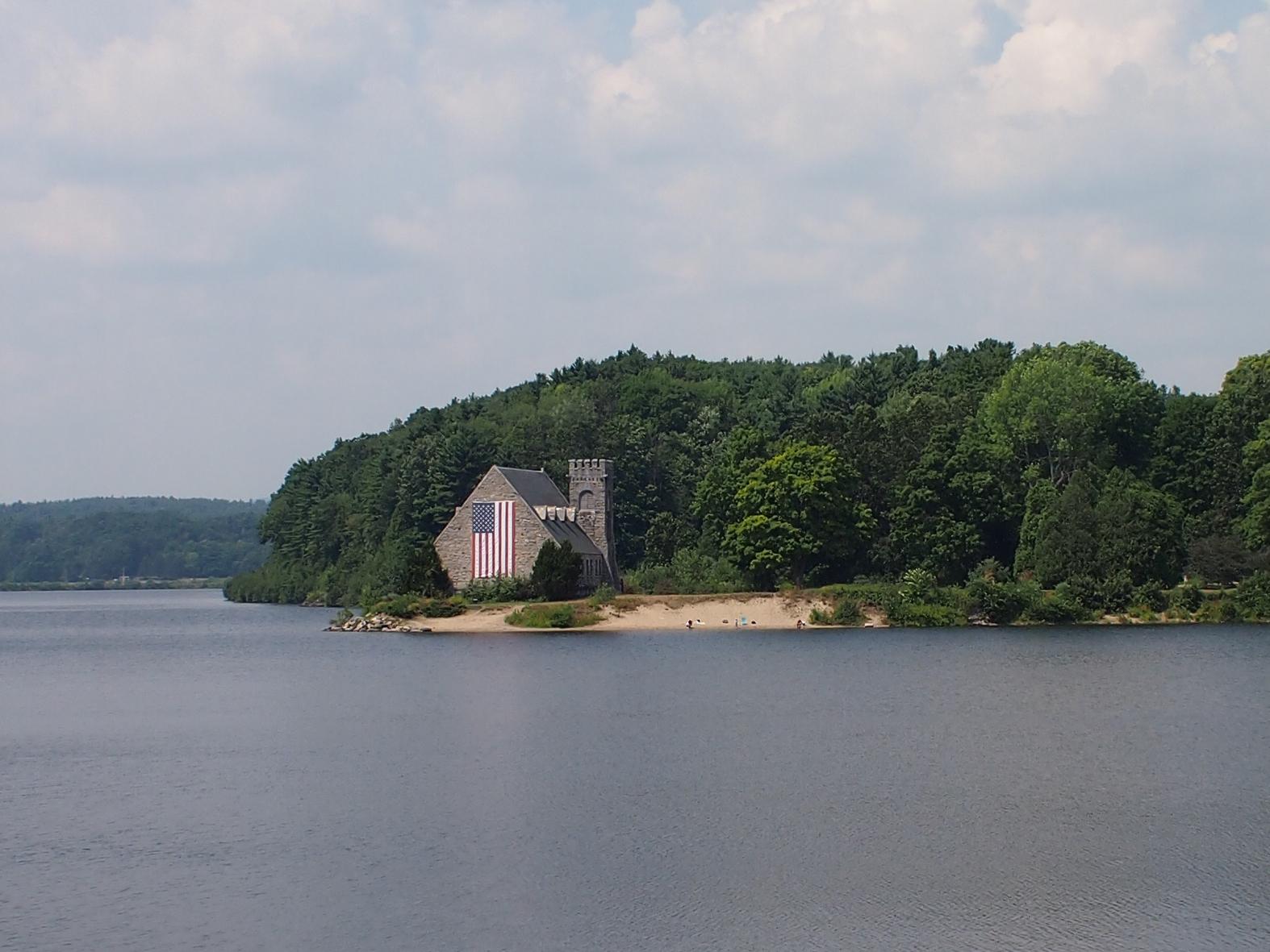Stone building with flag on sandy beach by water and green forest under cloudy sky.