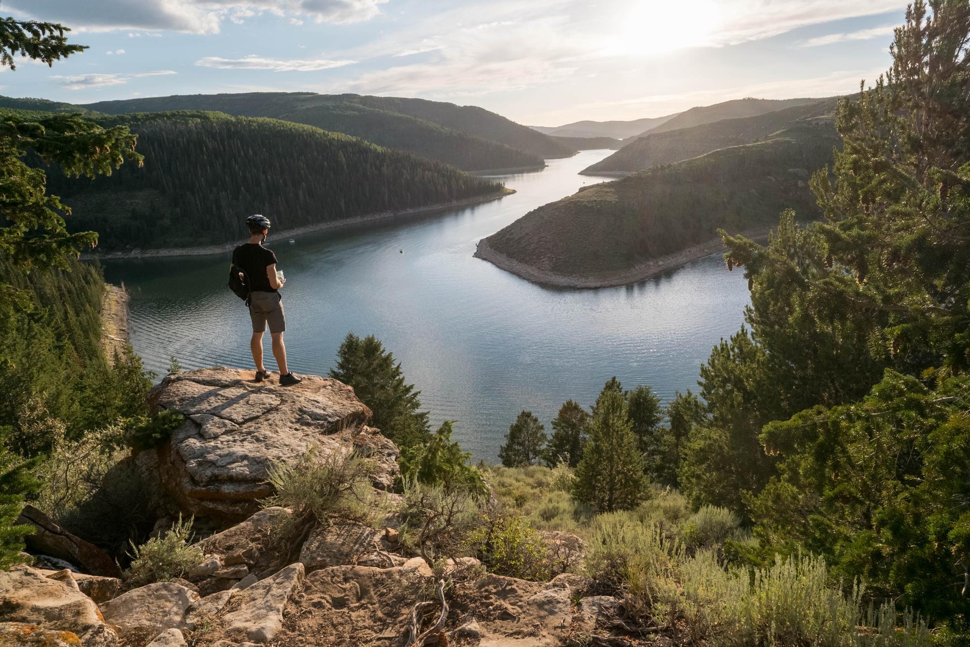Person on a rock overlooks a winding lake and forested hills at sunset.