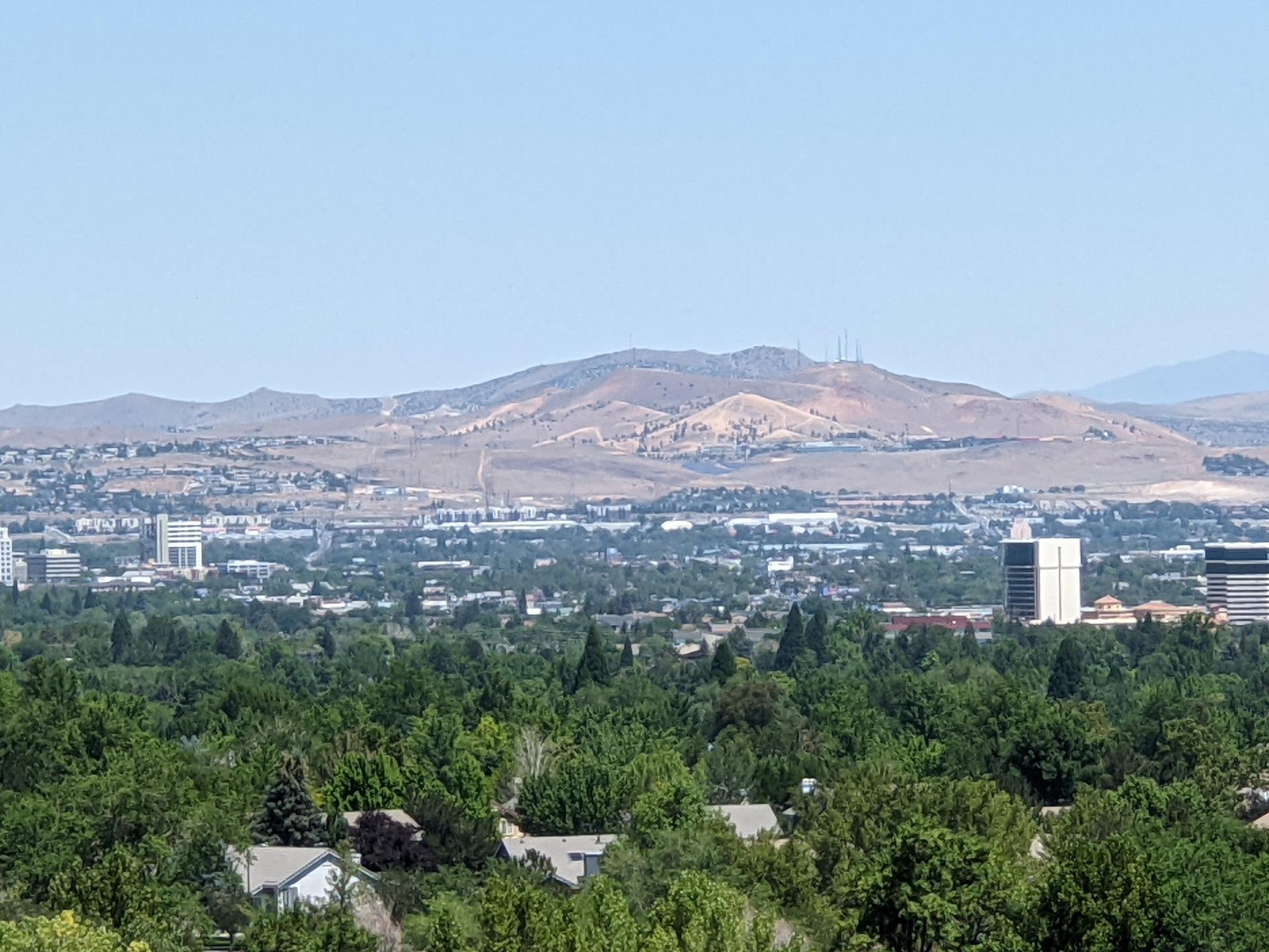 Expansive view of a city with lush trees, buildings, and arid mountains under a clear sky.