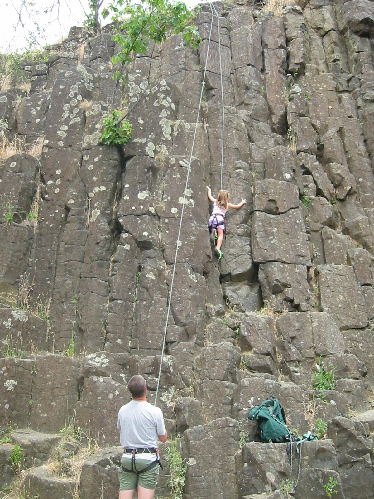 A person rock climbing a tall, dark cliff while another person belays below.