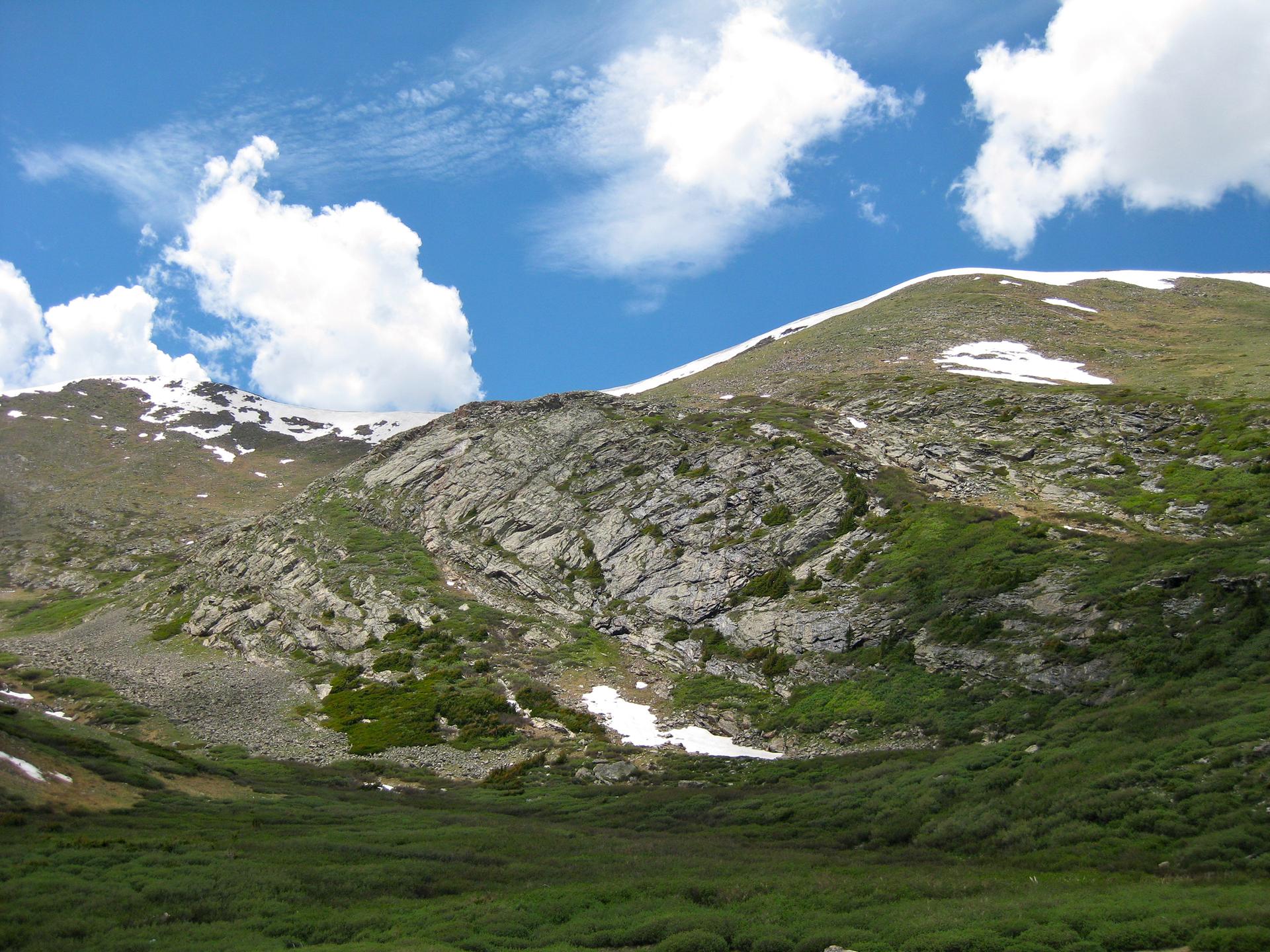 Rocky, snow-dusted mountainside with green shrubs under a bright blue sky.