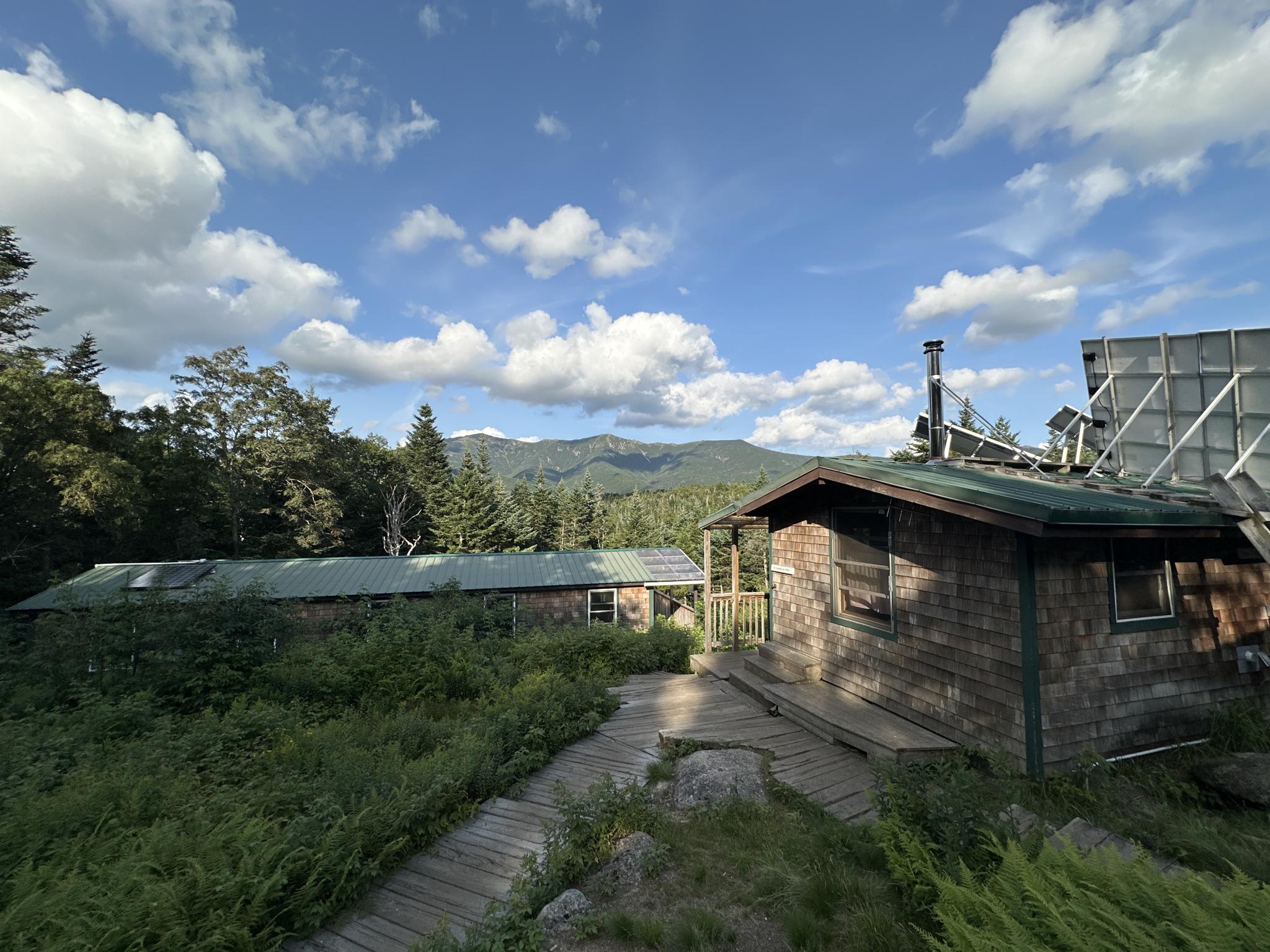 Two rustic cabins with a wooden walkway, surrounded by green foliage and mountains.