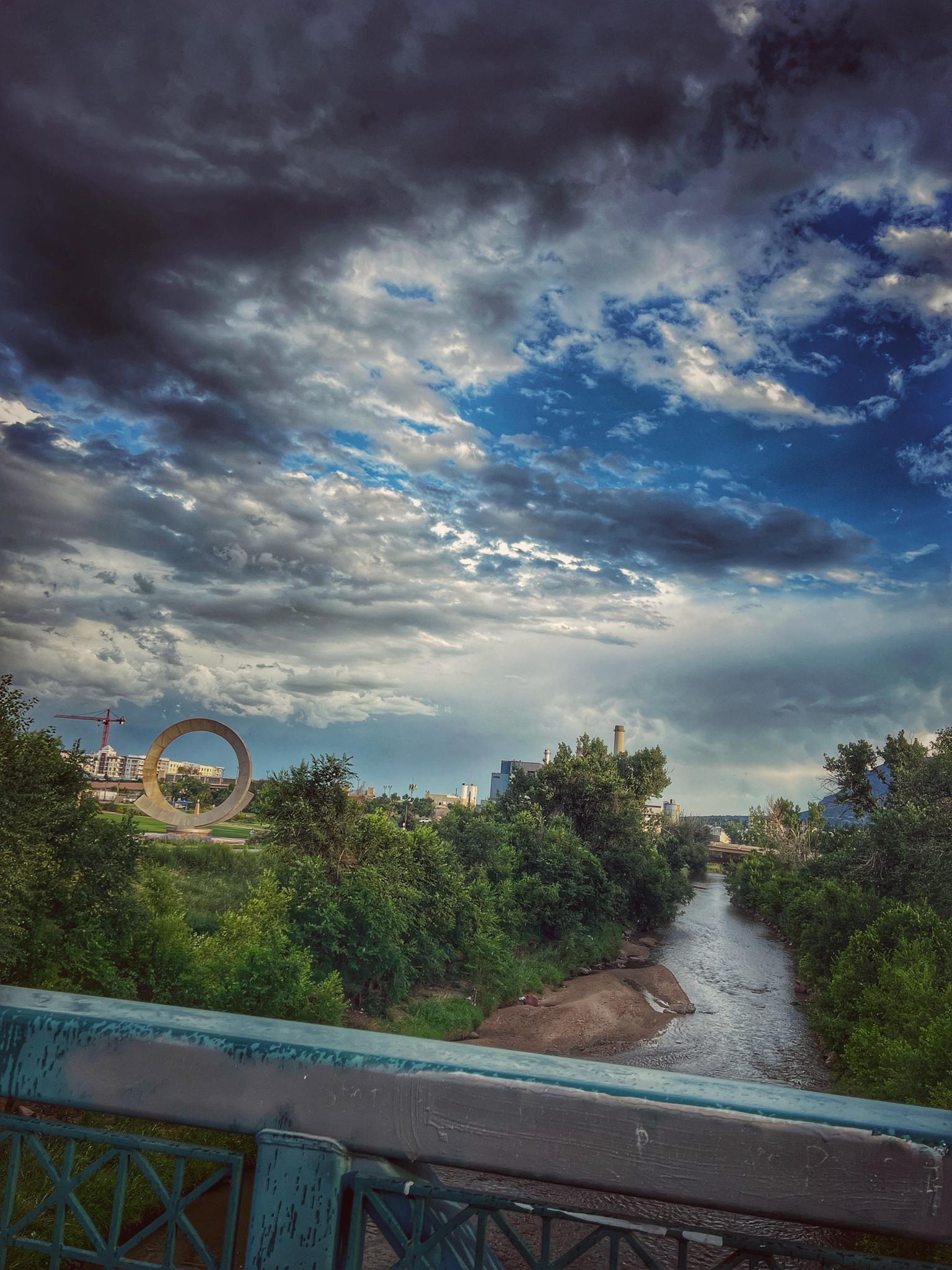 River flowing under a bridge with dramatic cloudy sky and distant structures.
