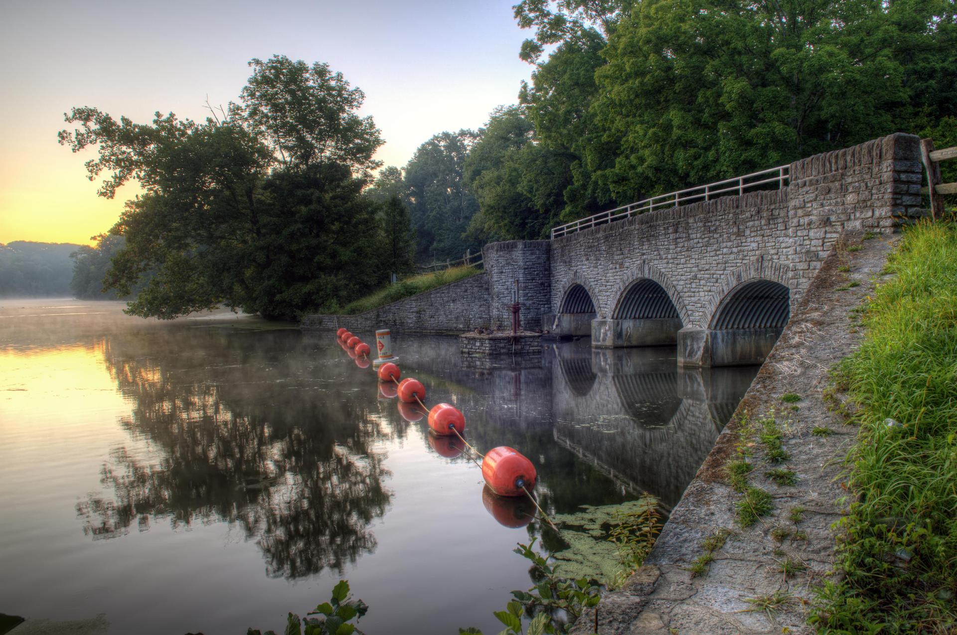 Stone arch bridge over misty water with orange buoys at sunrise.