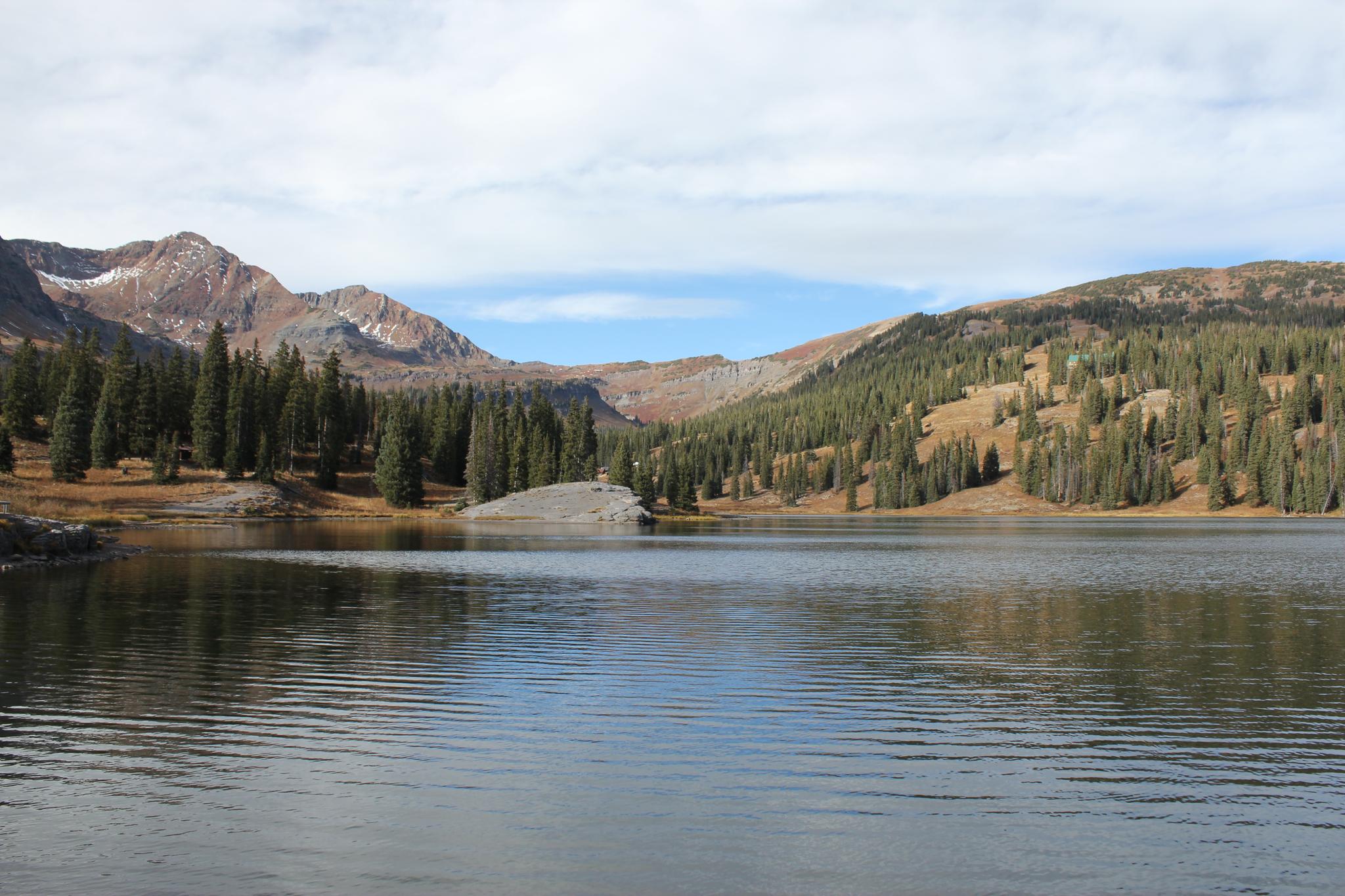 Calm mountain lake reflecting forested slopes and rocky peaks under a bright sky.