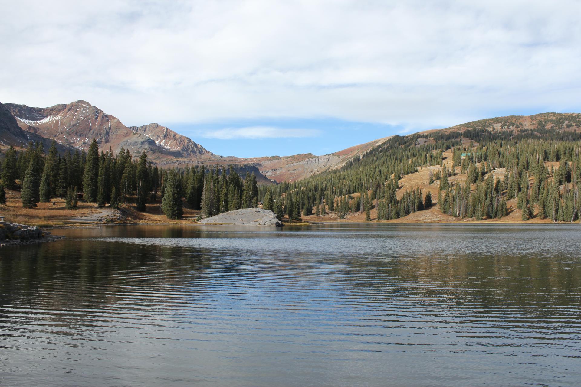 Calm mountain lake reflecting forested slopes and rocky peaks under a bright sky.