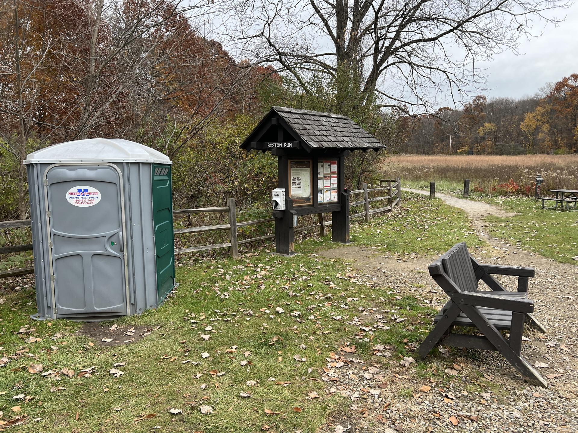 Portable toilet, information board, and bench on a grassy path under cloudy skies.