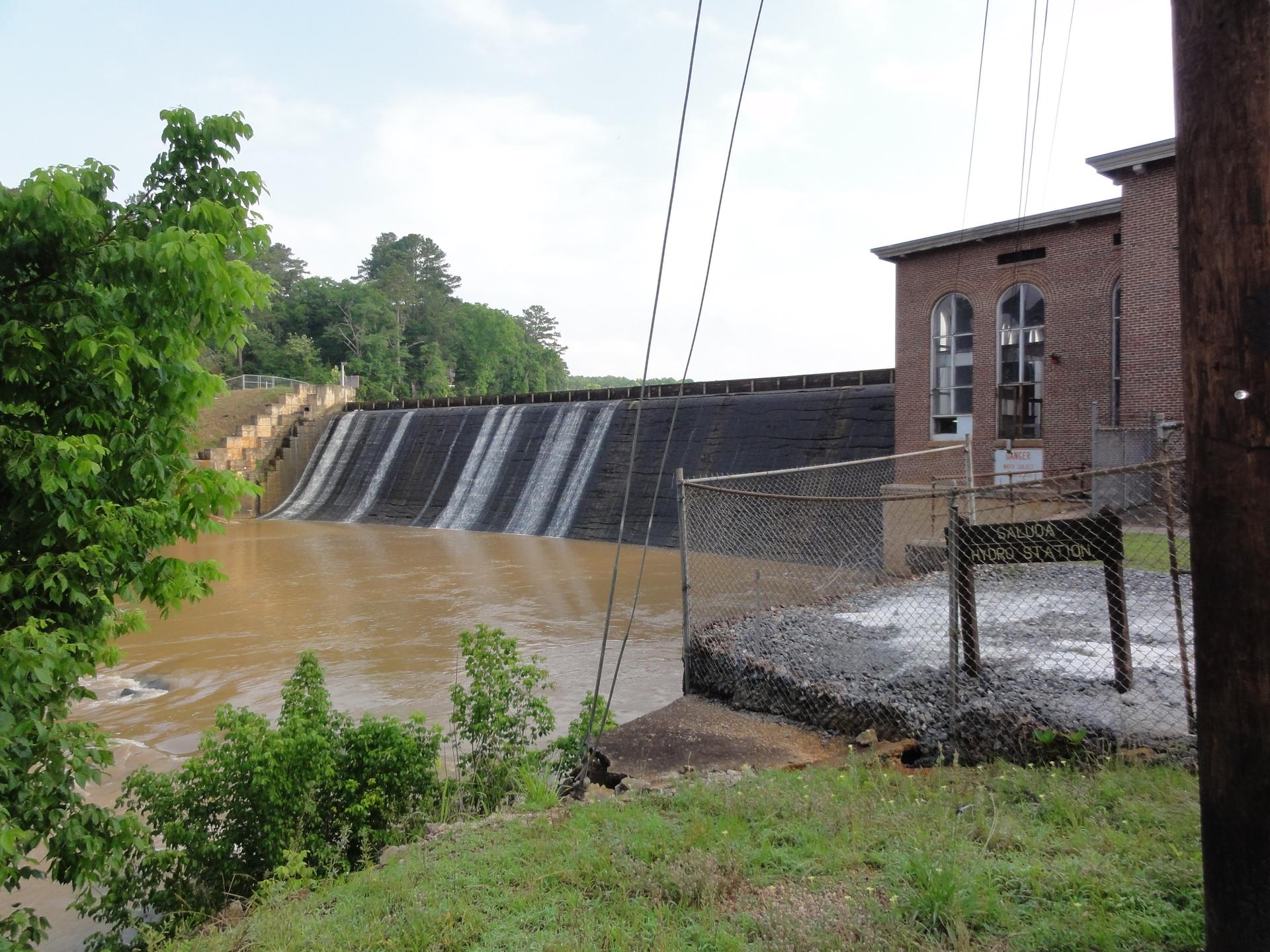 Muddy water flows over a dam next to a brick building under an overcast sky.