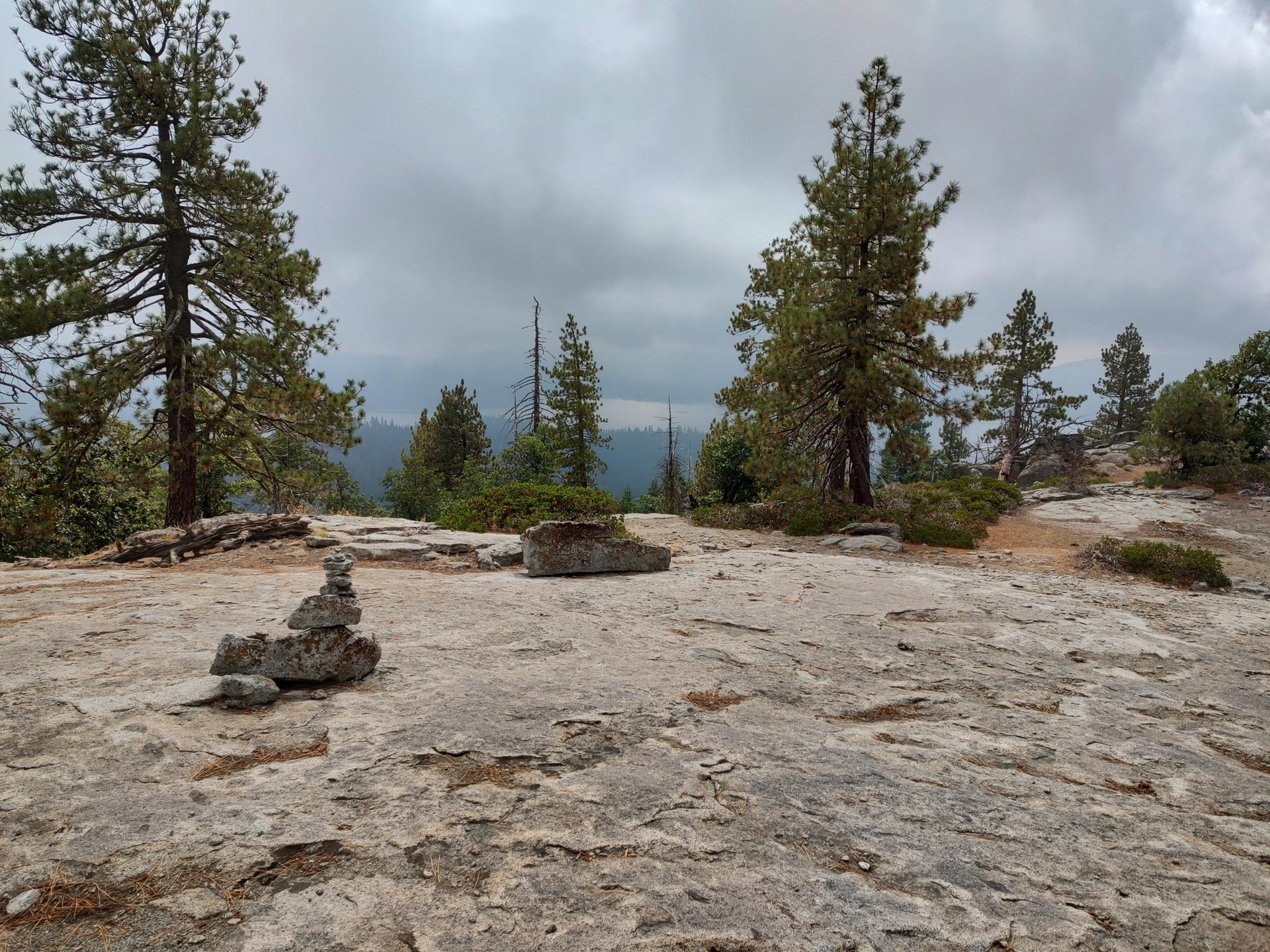 A rock cairn on a rocky outcrop with pine trees under a cloudy sky.