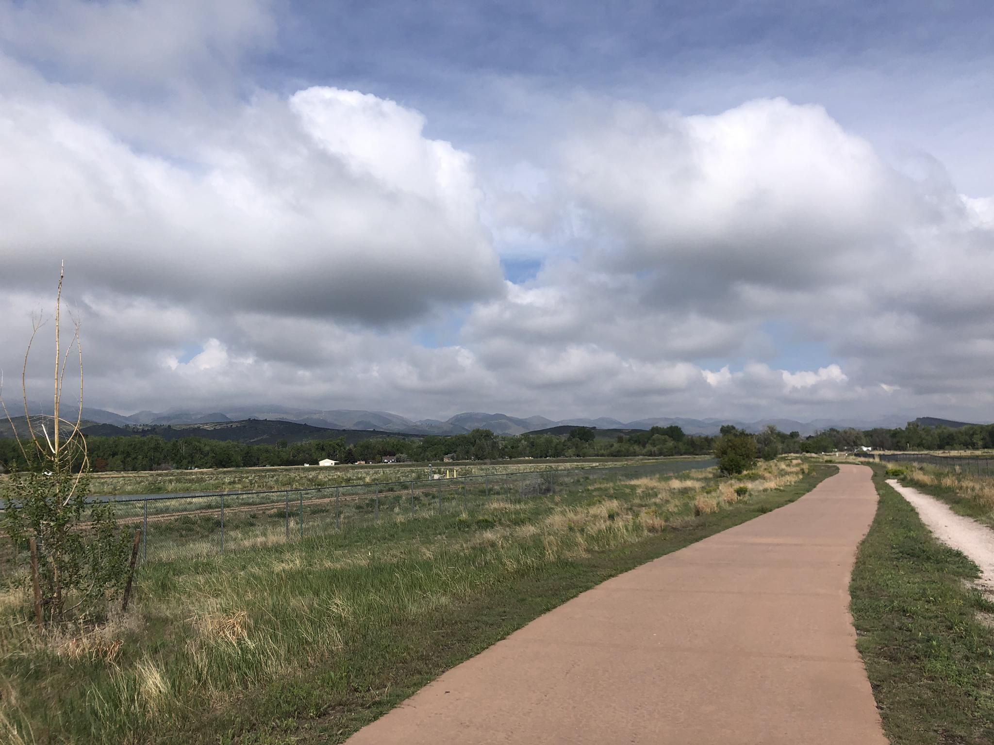 Paved path through a grassy field with distant mountains under a cloudy sky. - Poudre River Trail