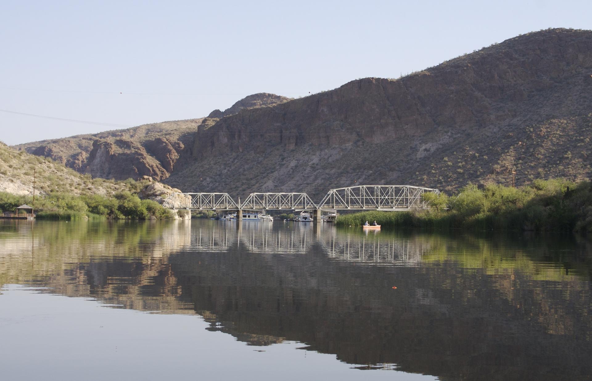 Long metal bridge spanning a calm river with distant mountains.