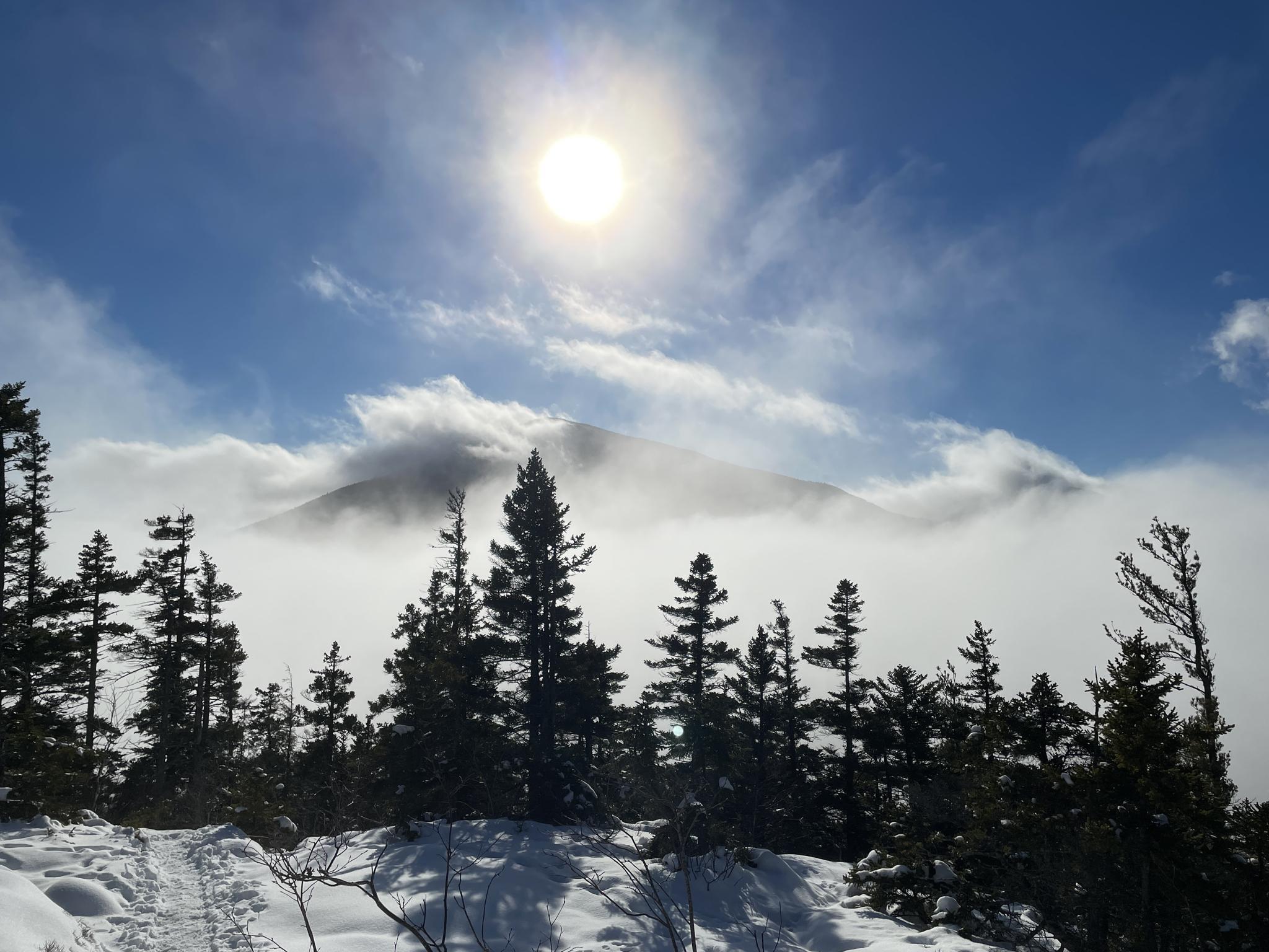 Bright sun over a snowy mountain peak shrouded in clouds, with evergreen trees.