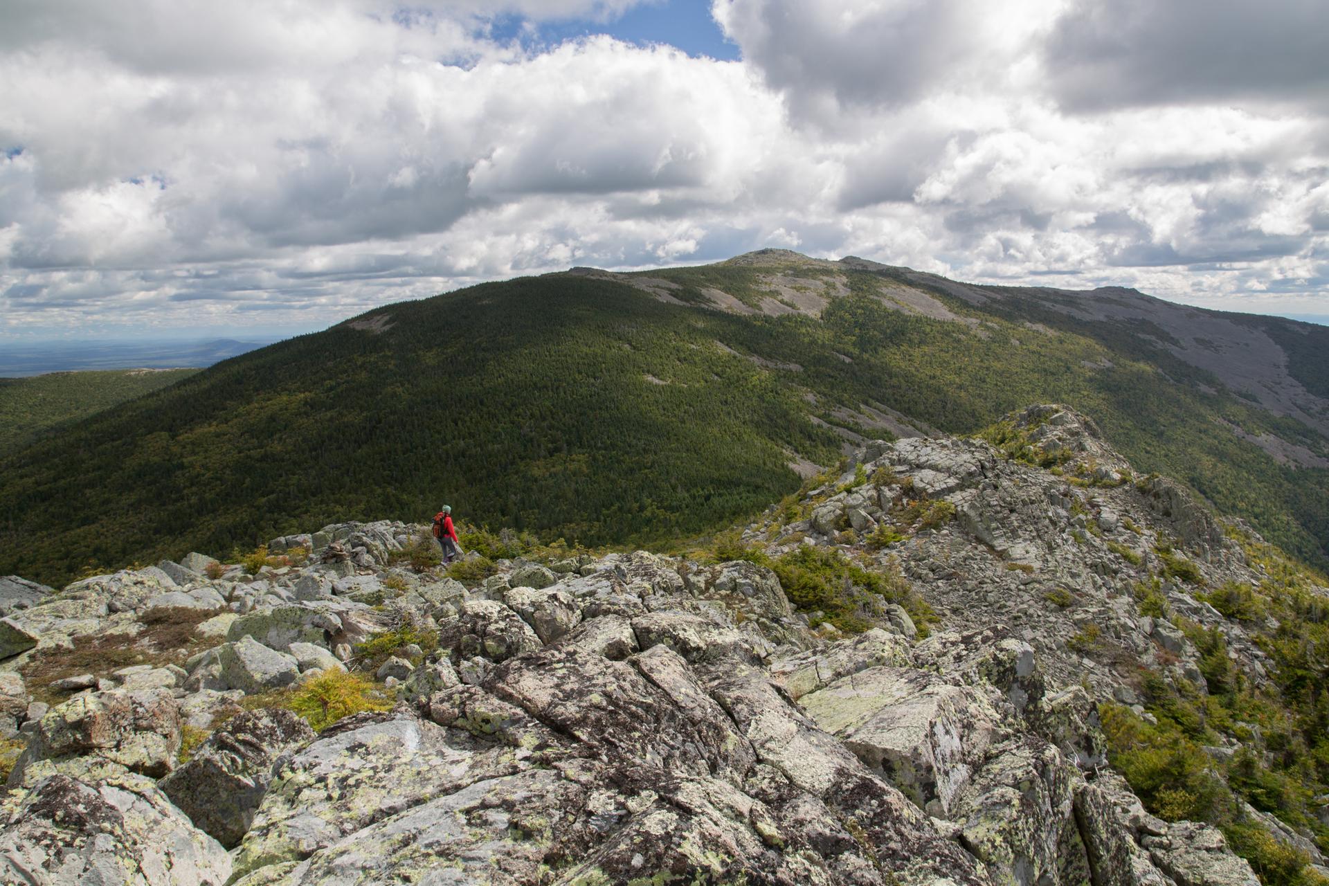 A hiker stands on a rocky mountain ridge under a cloudy sky.