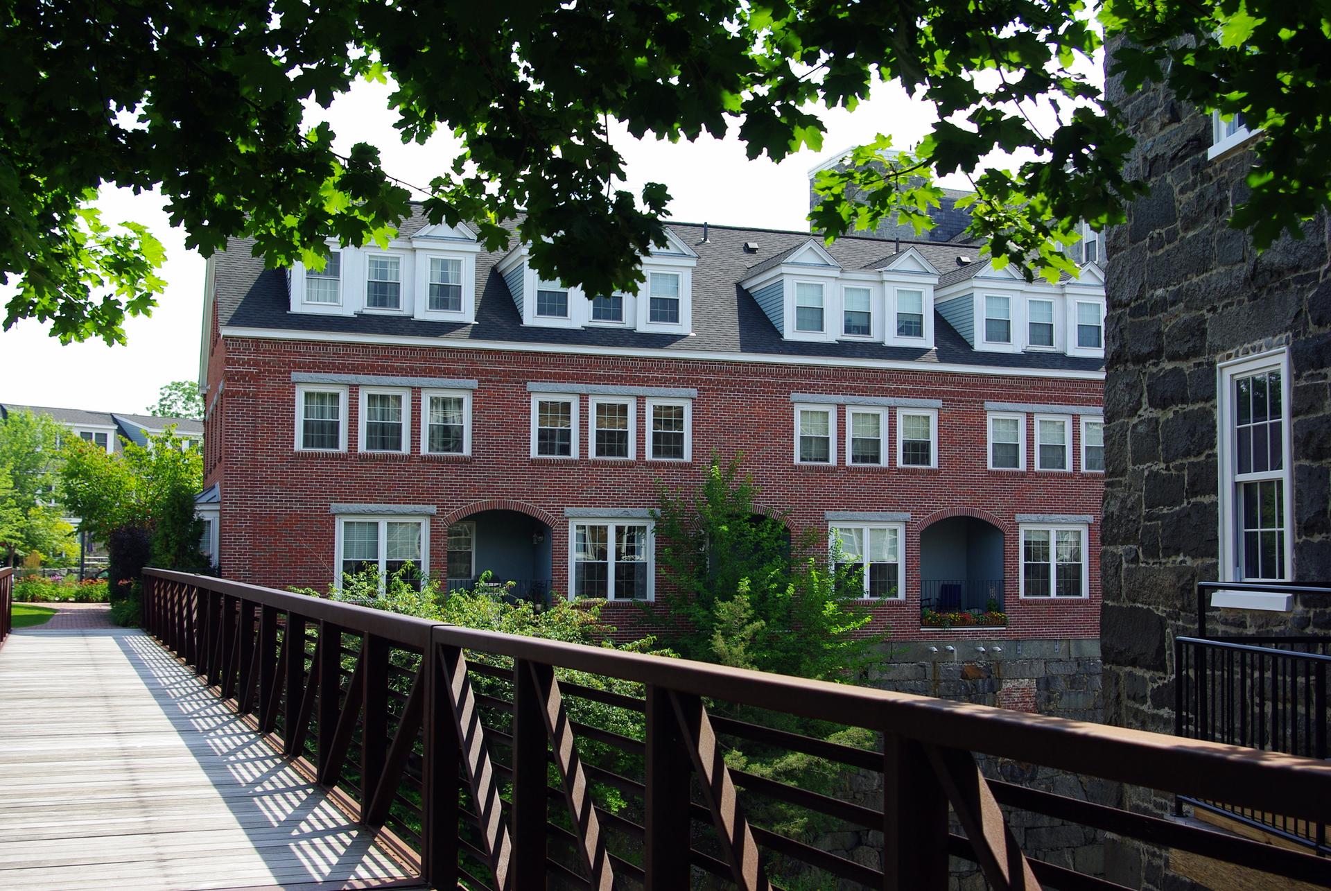 Red brick building with dormers viewed from a wooden bridge on a sunny day.