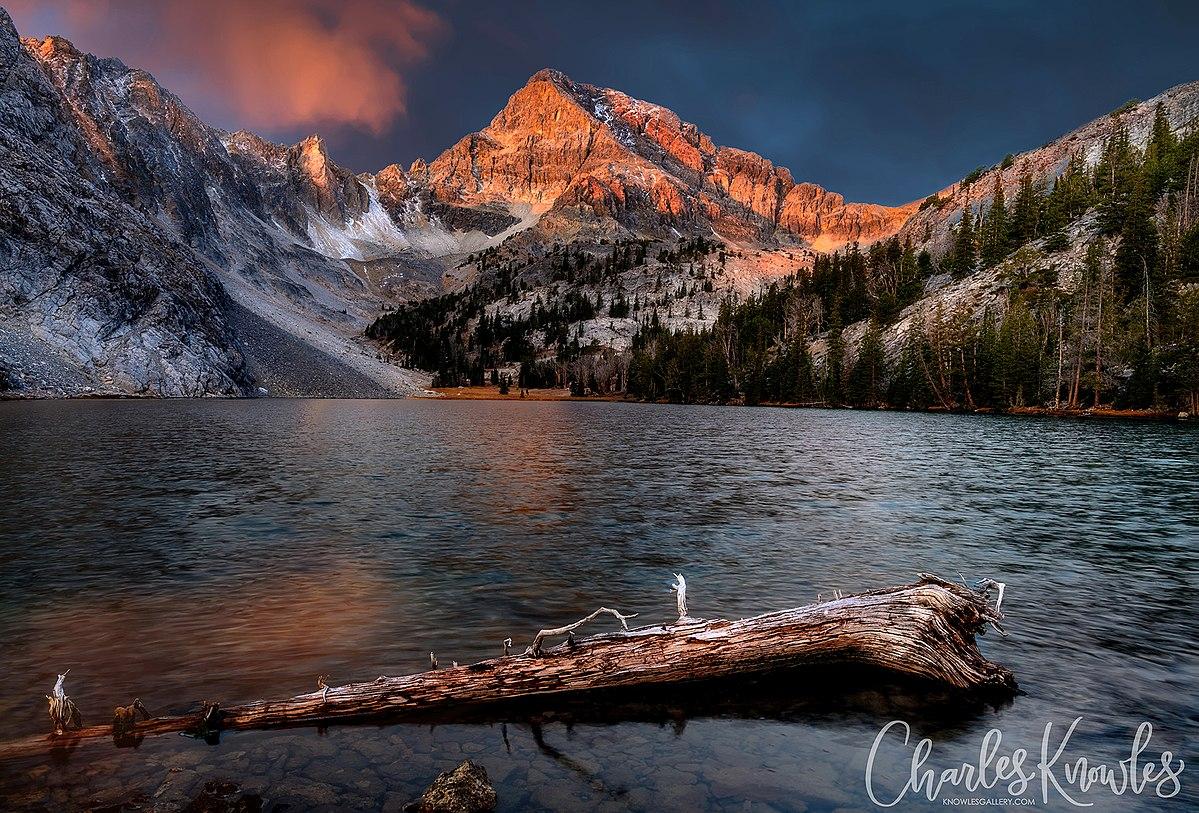 Mountain lake at sunset with golden peaks, dark sky, and a log in the water.