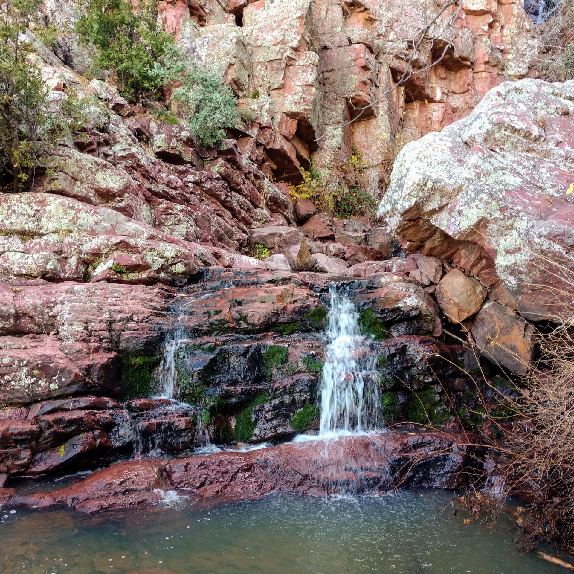 Small waterfalls cascade over red rocks into a clear pool.