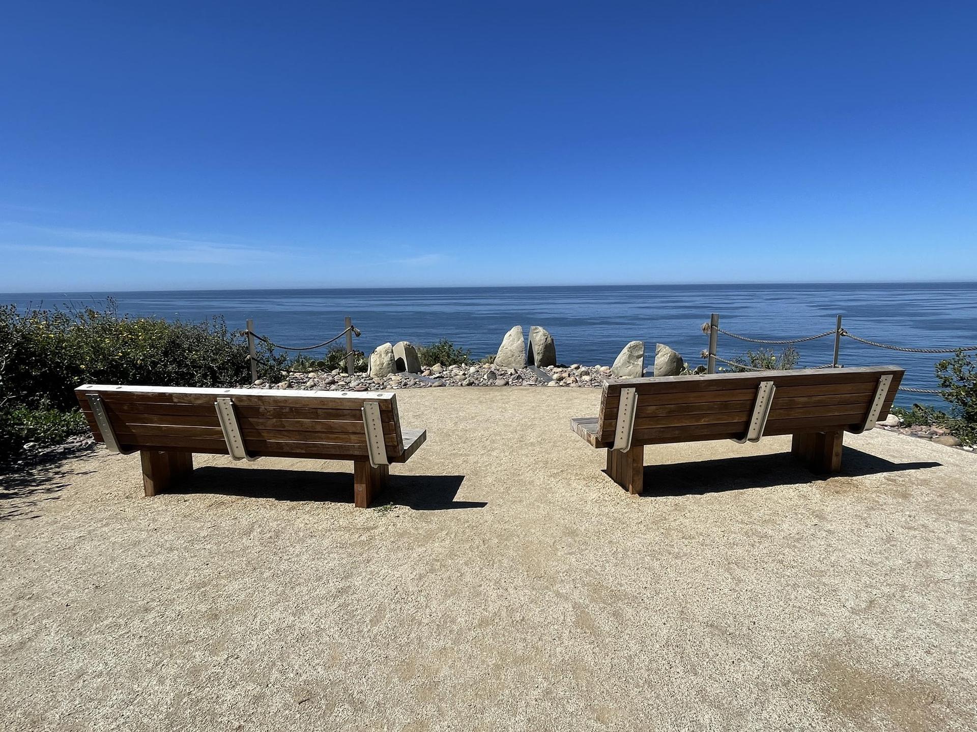 Two wooden benches face the calm ocean under a bright blue sky.