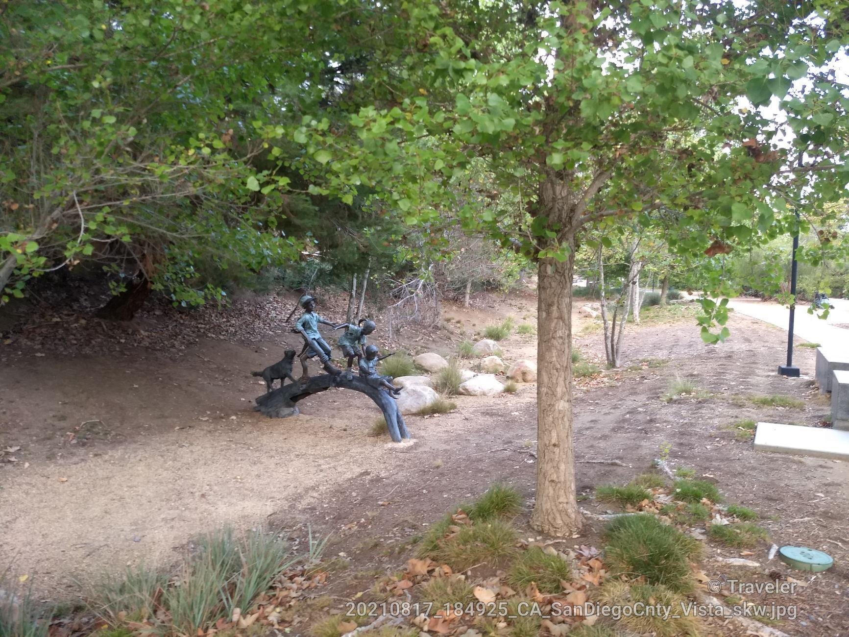 Bronze sculpture of children and a dog on a log in a wooded park.