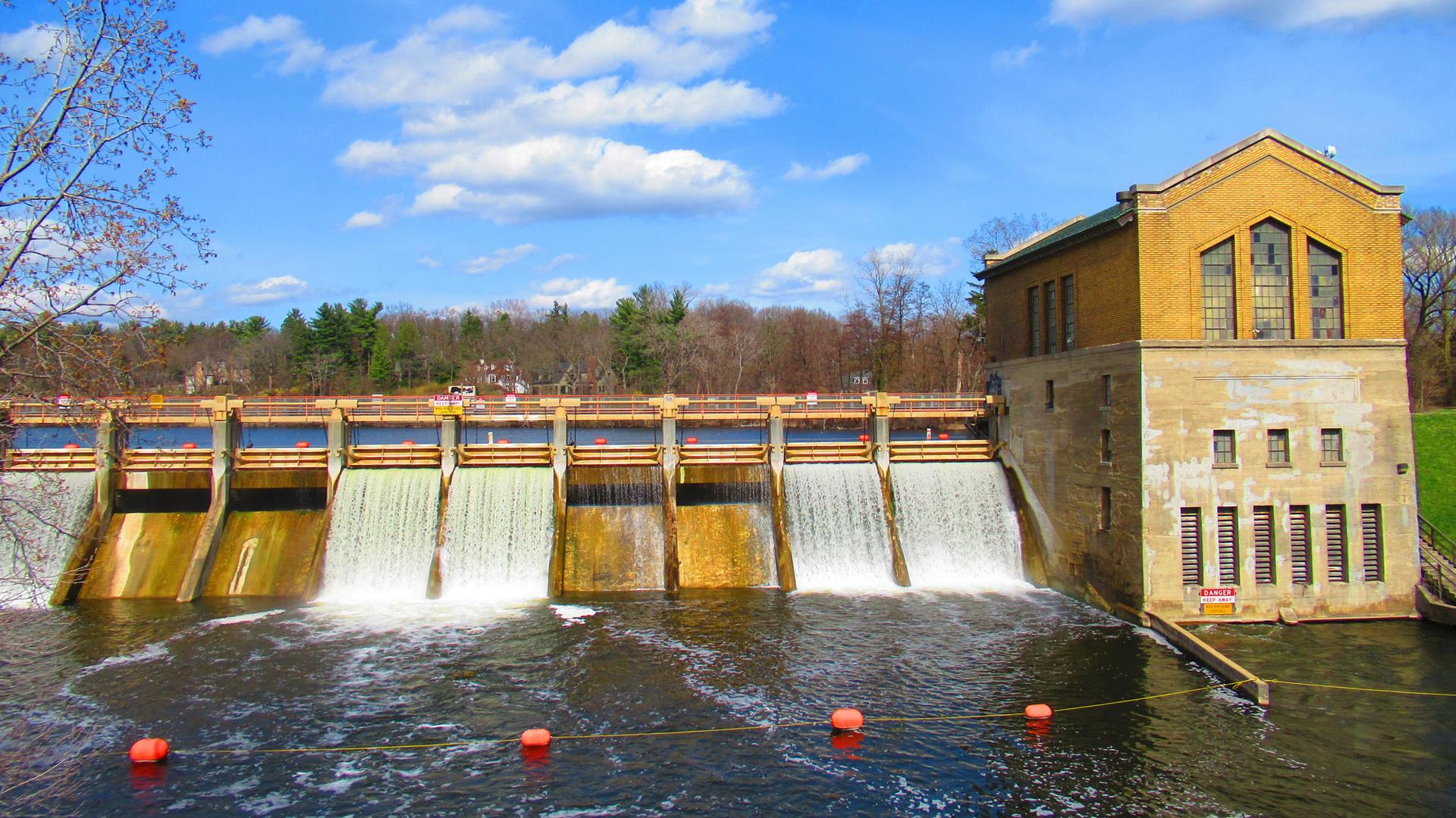 A dam with water flowing over gates, next to a large brick building.