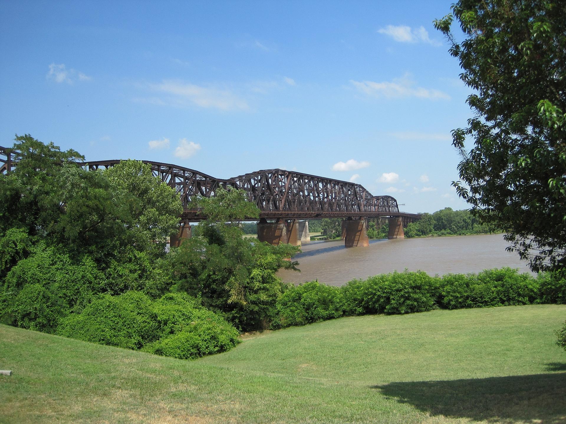 Large metal bridge over a wide river, surrounded by green trees and grass.