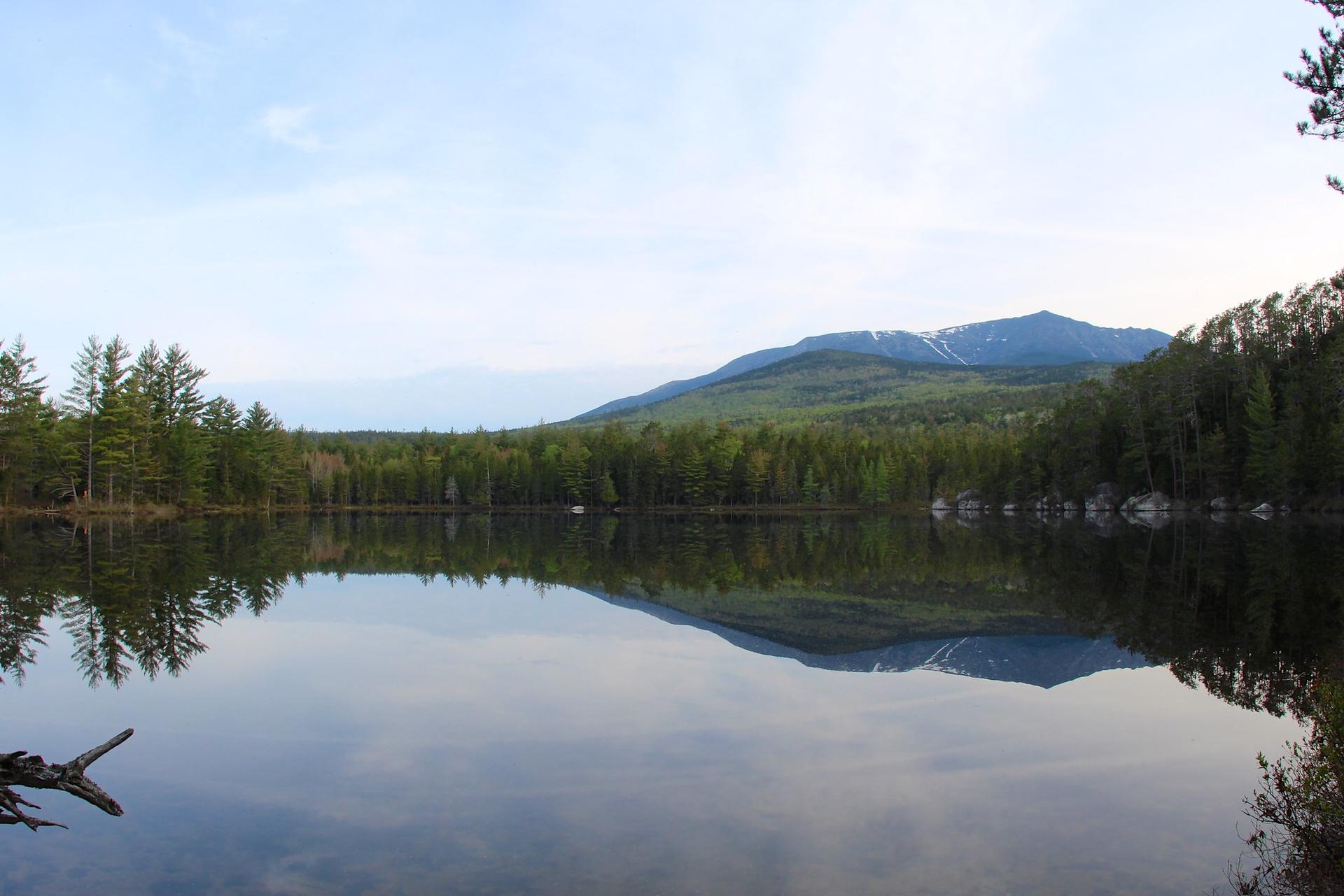 Calm lake reflecting forested mountains with snow under a clear sky.